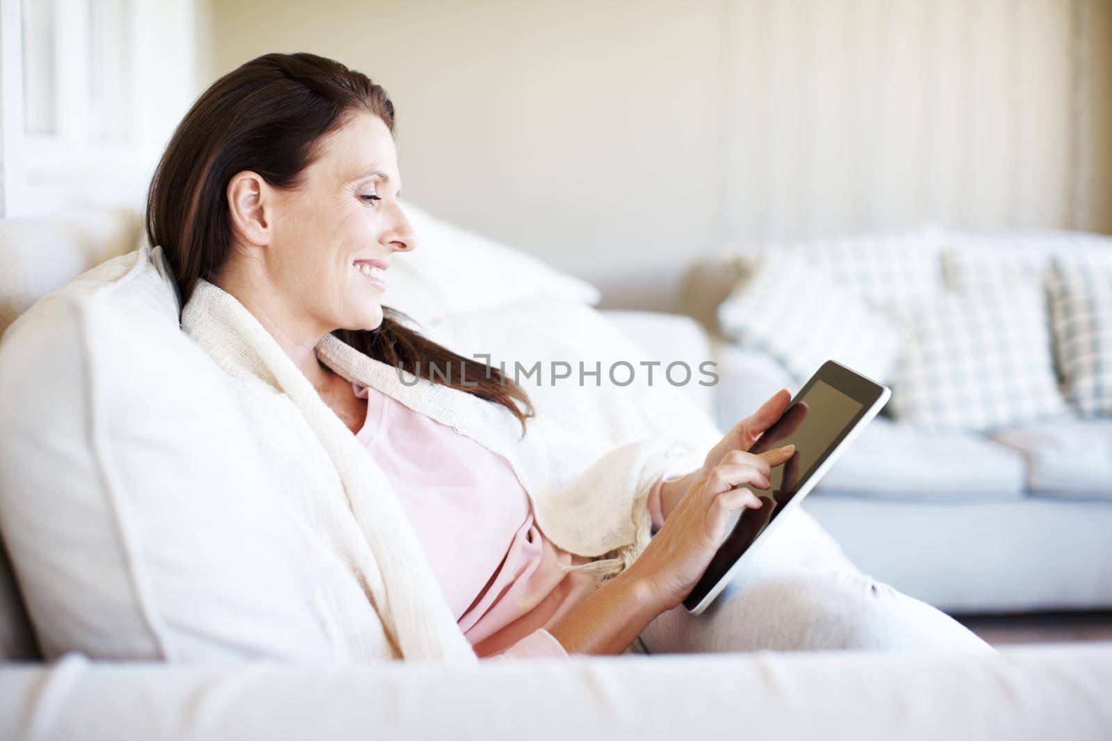 Reading a romance novel on her e-reader. An attractive woman using a digital tablet while sitting on a sofa. by YuriArcurs