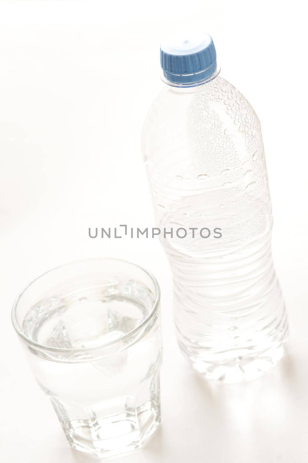 Bottled water served in a glass tumbler with the half full capped unlabeled plastic bottle alongside, high angle tilted view