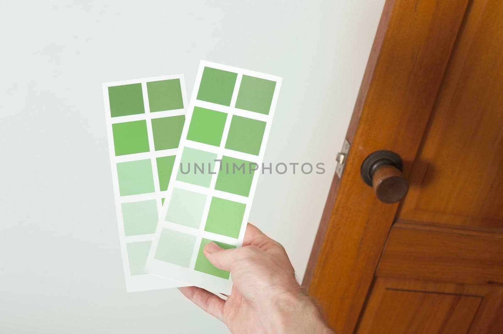Man holding up cards of green paint swatches against a white wall in his house as he makes a decision on colors to redecorate the interior