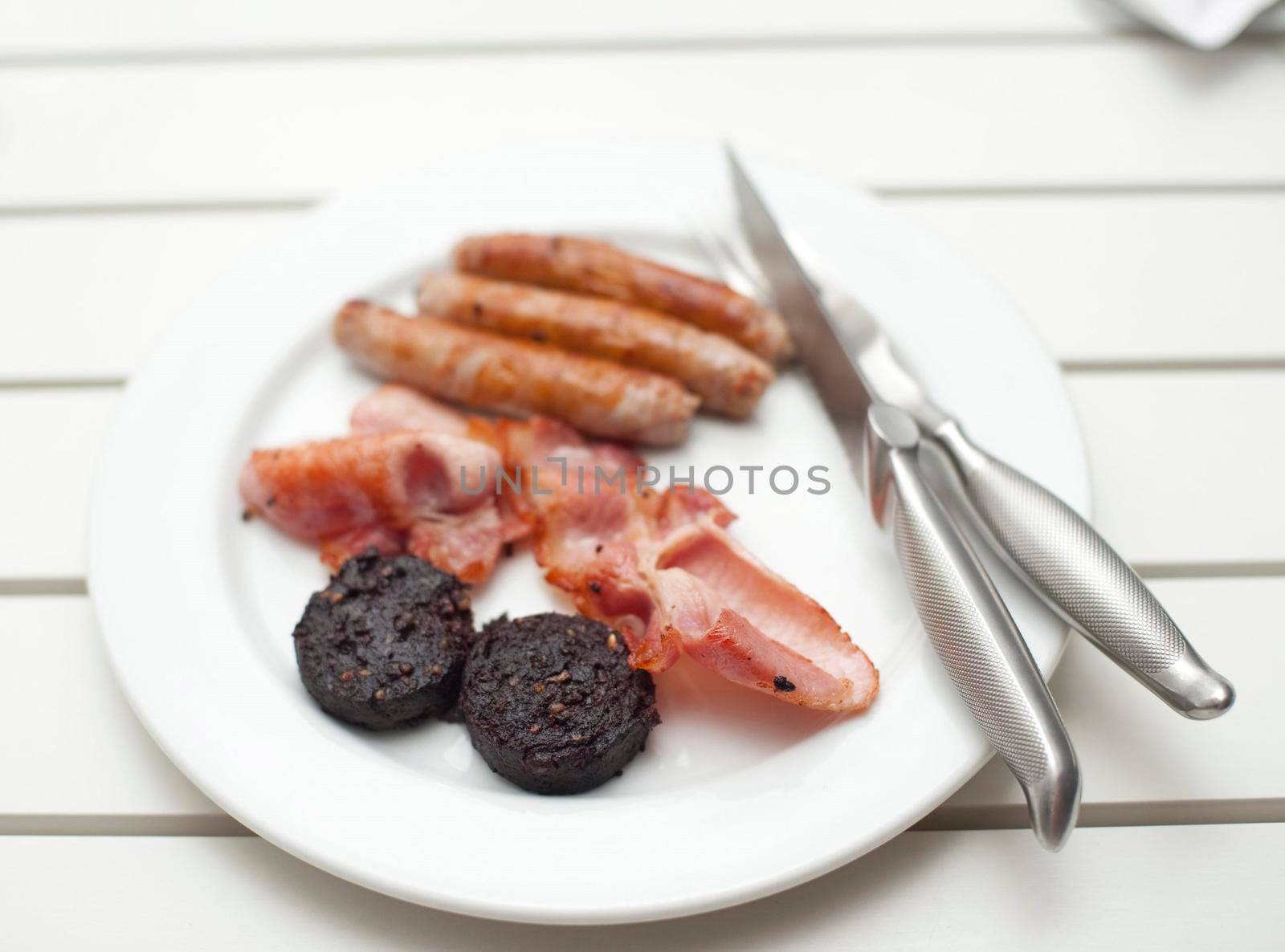 Fried breakfast with mushrooms, crispy bacon and pork sausages served on a white plate on a painted white slatted table with shallow dof
