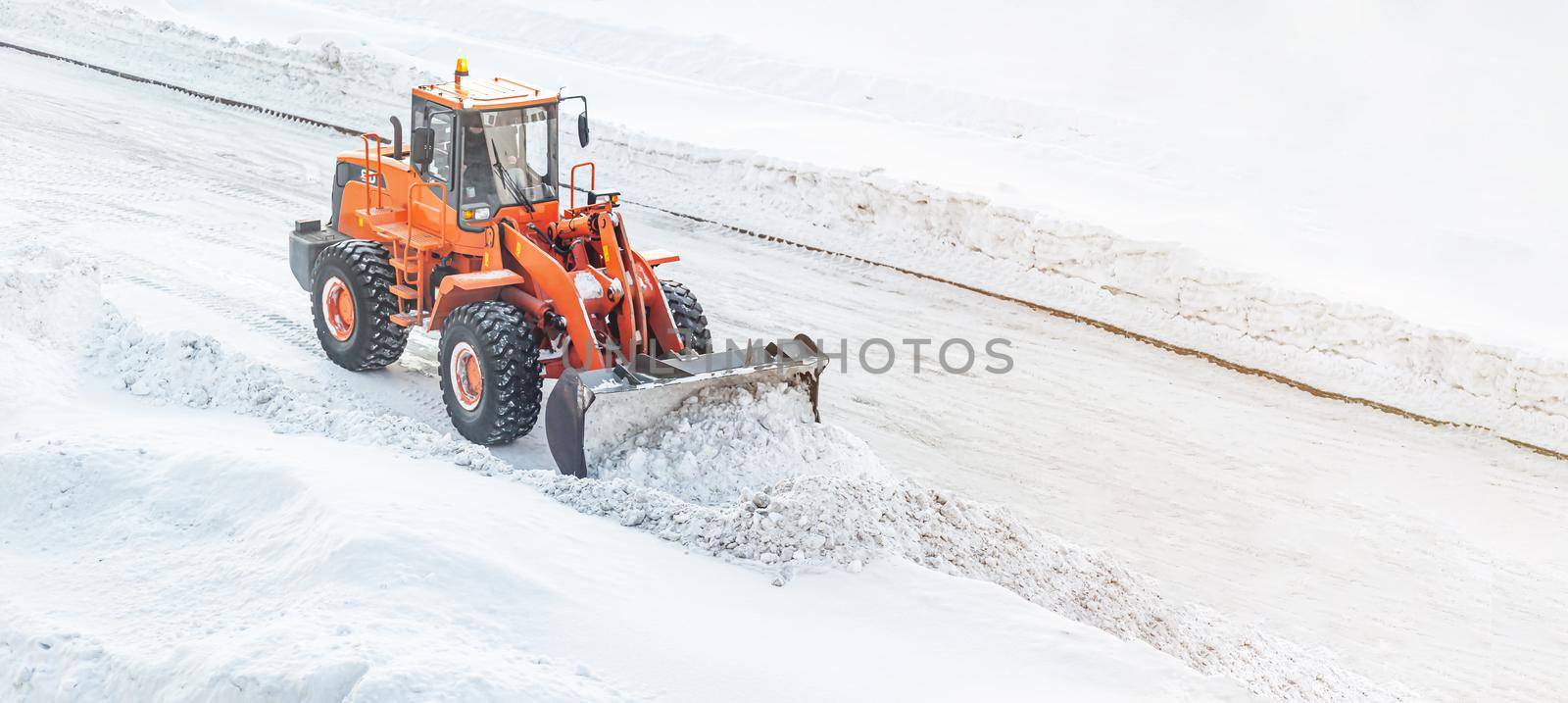 A large orange tractor removes snow from the road and clears the sidewalk. Cleaning and clearing roads in the city from snow in winter. Snow removal after snowfalls and blizzards.