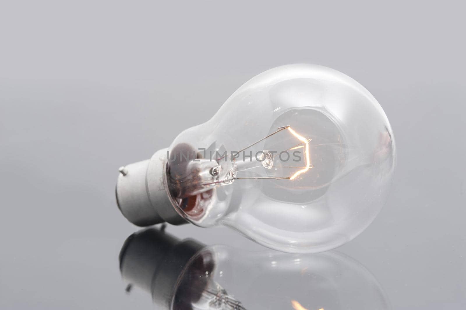 Domestic light bulb with a bayonet fitting and a glowing filament on a reflective grey surface for power and energy or bright ideas concepts