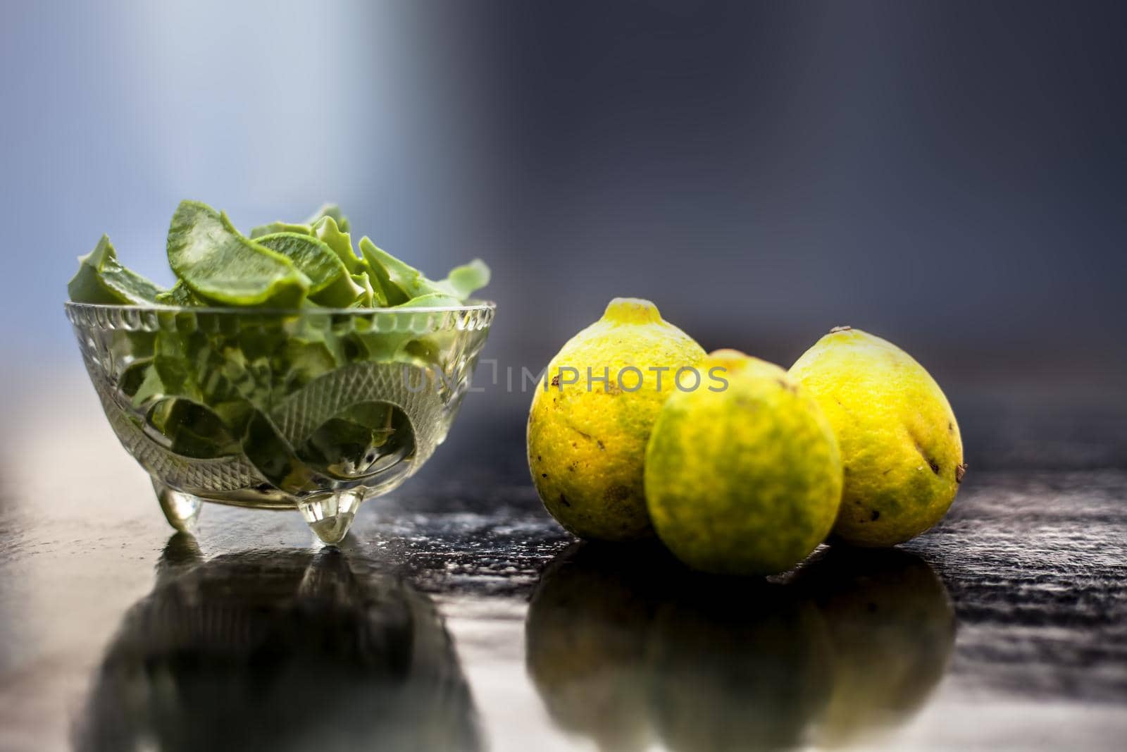 Raw cut aloevera or aloe vera gel in a glass bowl and some fresh ripe lemons or limbu or nimbo on wooden surface along with their reflection on the surface.