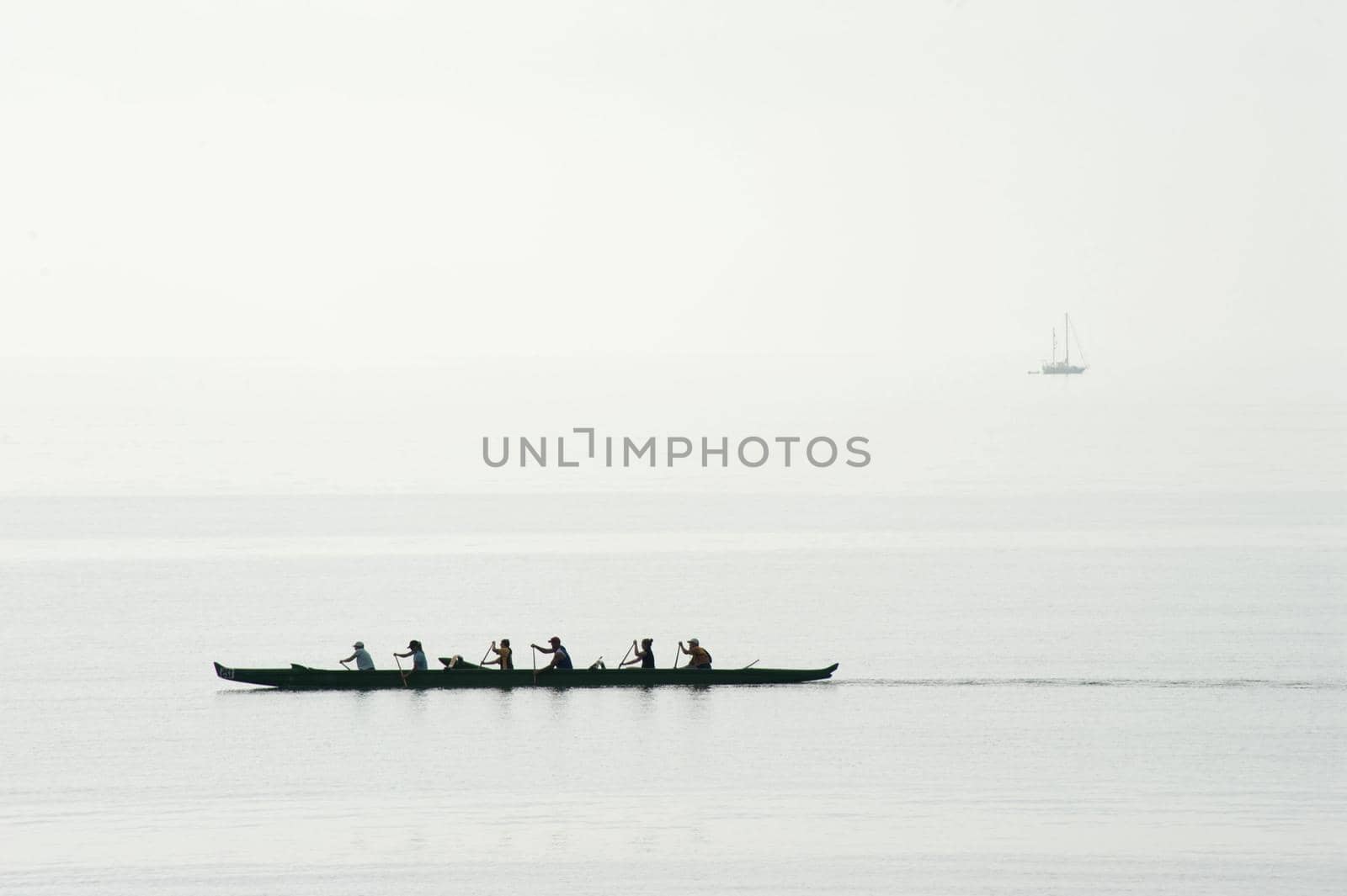 Six men team training for a race in their canoe paddling in unison on a calm ocean, silhouette against a misty sea
