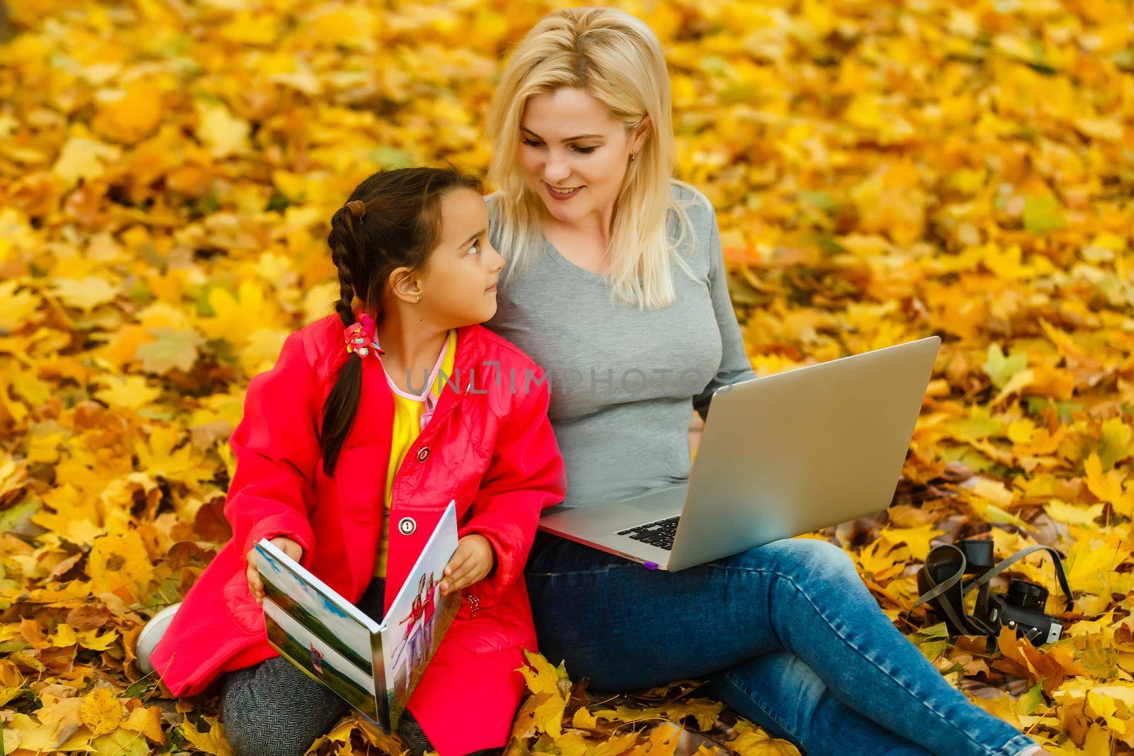 Happy smiling mother and child sits together looking on book or tablet pc in autumn park