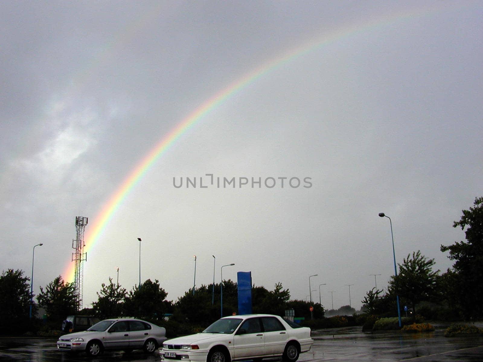 Colorful Rainbow Arching Over Rain Soaked Parking Lot in Cloudy Sky in Aftermath of Rain Storm