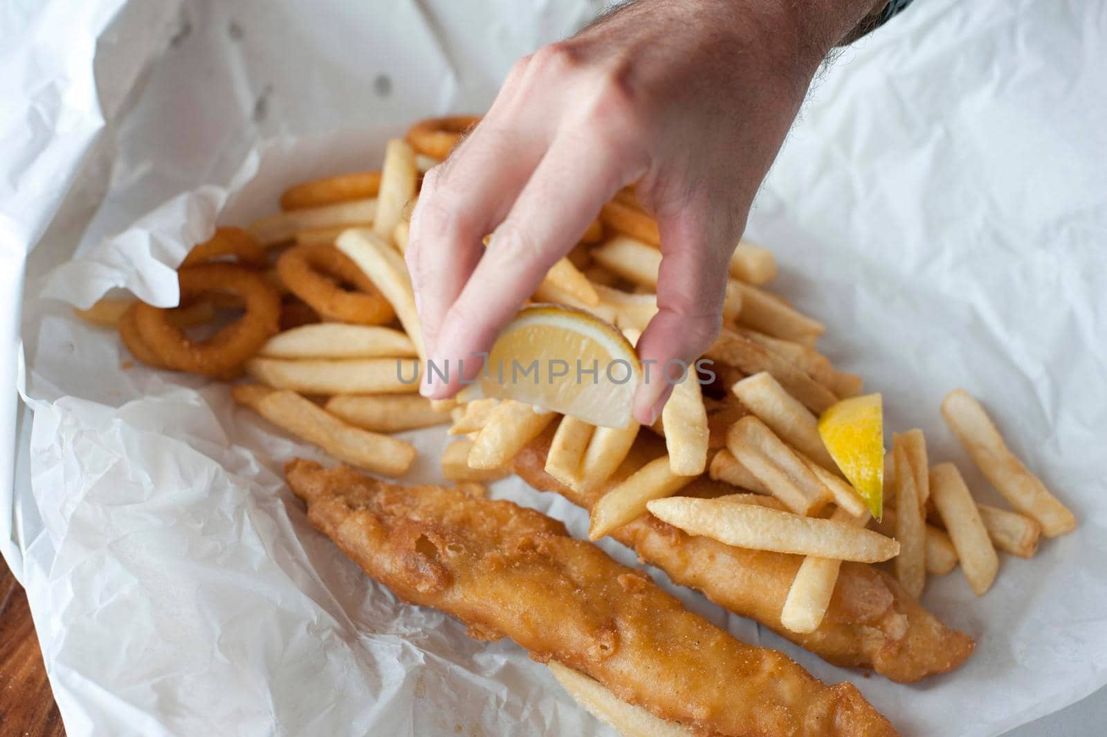 Hand squeezing lemon over takeaway fried fish fillets and calamari in batter with fried potato chips on crumpled paper