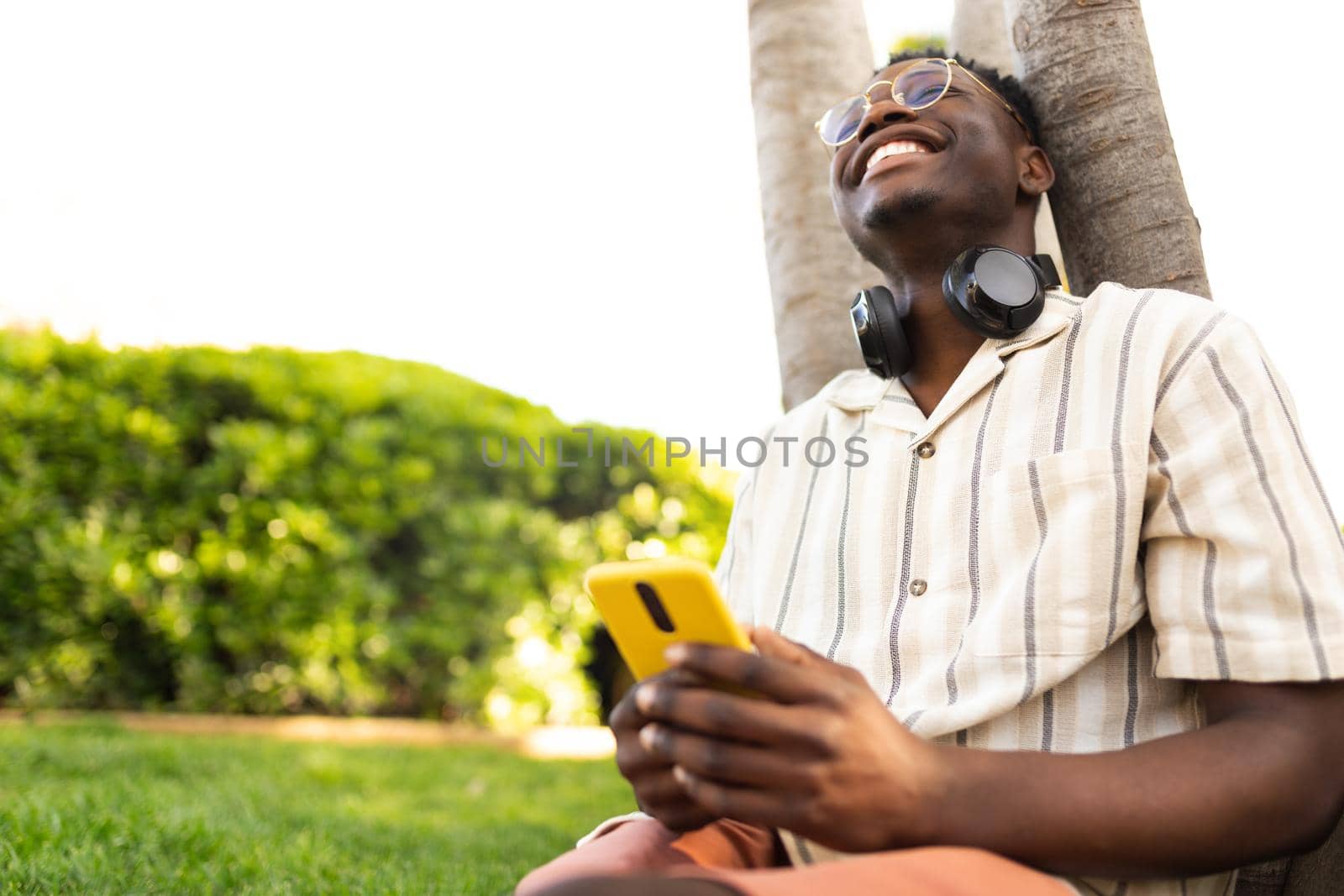 Happy black man relaxing outdoors using mobile phone. College student laughing on campus. Copy space. Lifestyle concept.