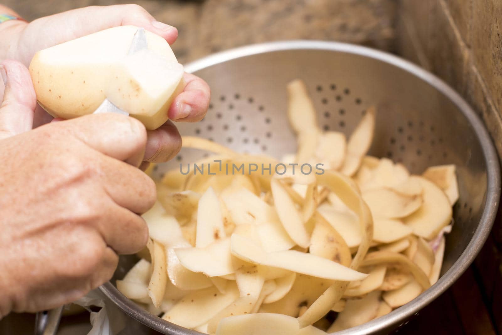 Man peeling fresh potatoes for cooking catching the peels in a metal colander, close up on his hands and the utensil