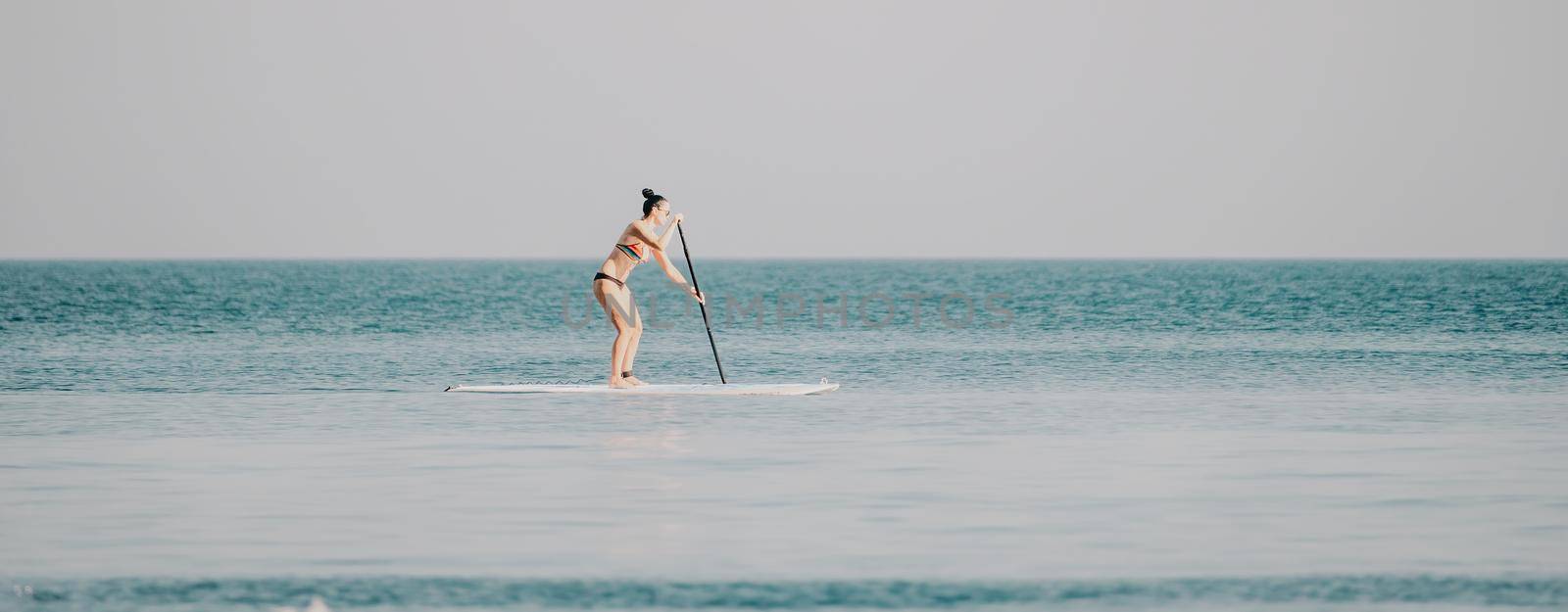 Silhouette of woman standing, surfing on SUP board, confident paddling through water surface. Idyllic sunset or sunrise. Sports active lifestyle at sea or river.