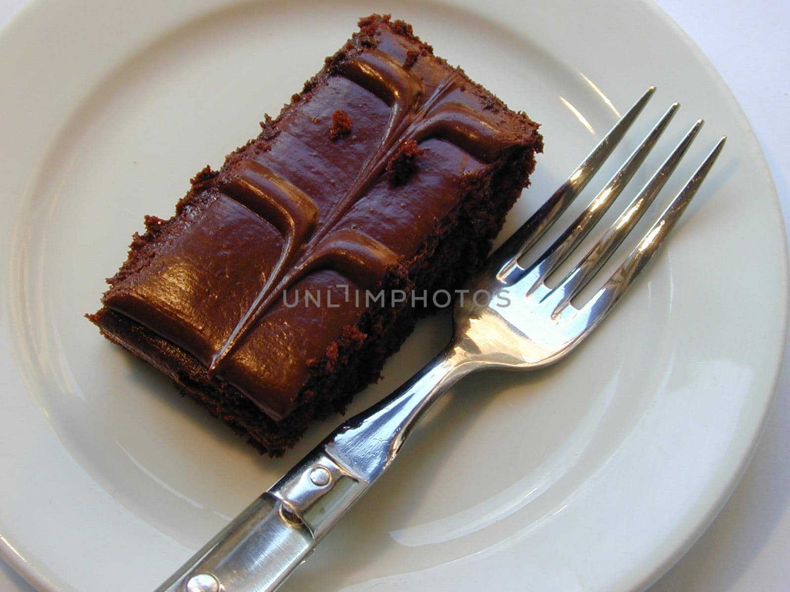 Delicious portion of rich glazed chocolate cake with a decorative patterned icing served on a white plate with a fork for tea, high angle view