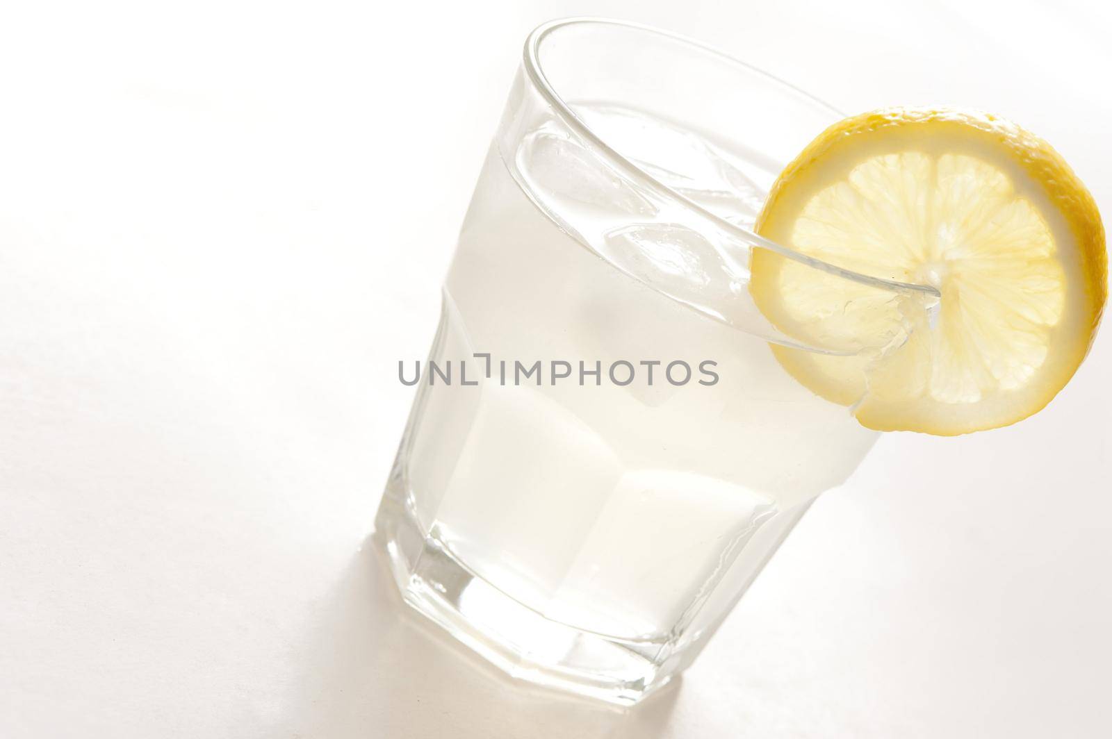 Long glass of clear spirits served with ice and garnished with a slice of fresh lemon, tilted angle close up on white