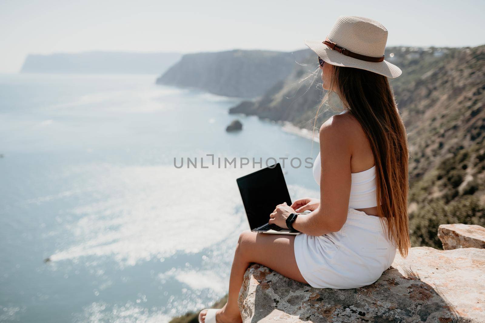 Successful business woman in yellow hat working on laptop by the sea. Pretty lady typing on computer at summer day outdoors. Freelance, travel and holidays concept.