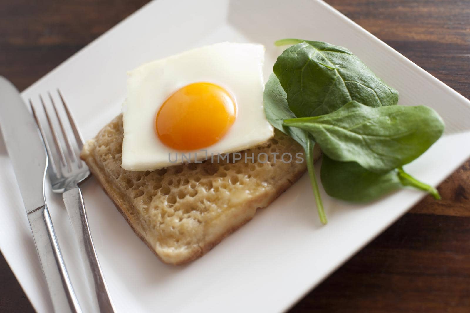 Close up of square egg sunny side up on a crumpet besides fresh spinach leaves and utensils on a white plate