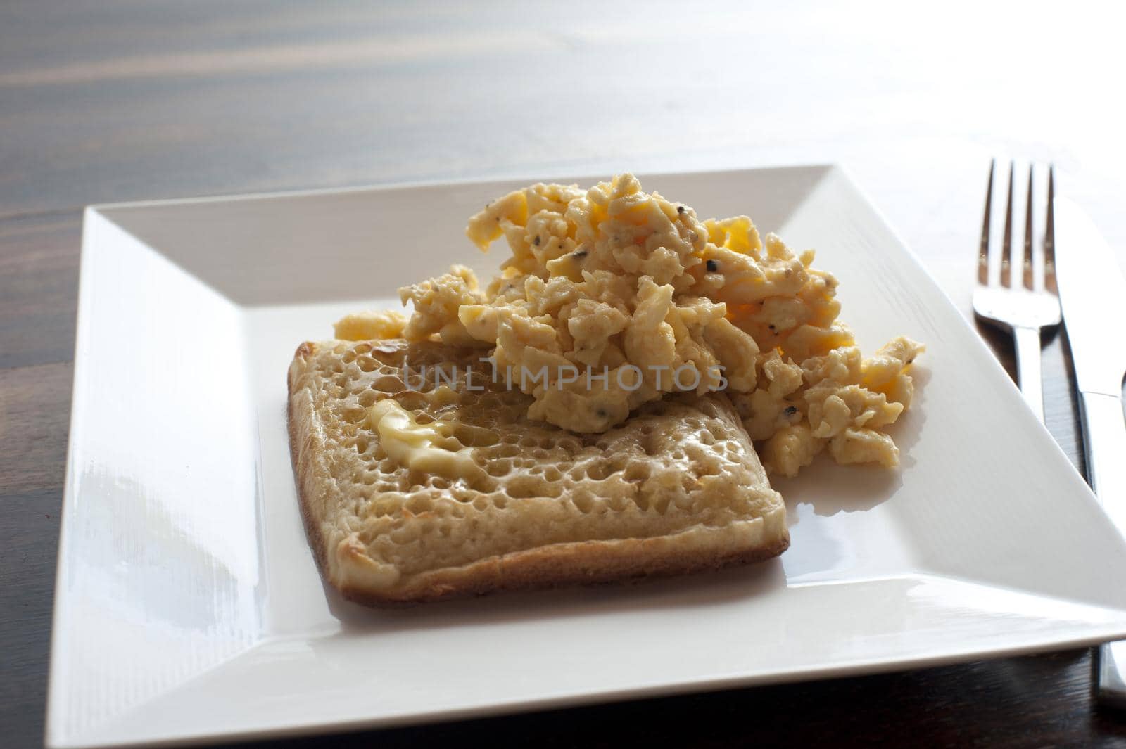 Single delicious buttered square crumpet and scrambled eggs in plate next to fork and knife on table
