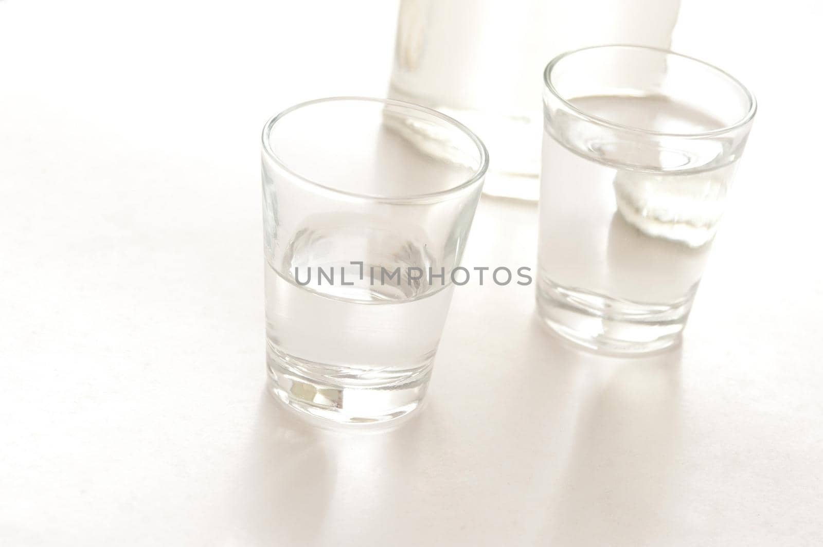 Two shot glasses with servings of white spirits, possibly neat vodka, with the base of a bottle of alcohol in the background