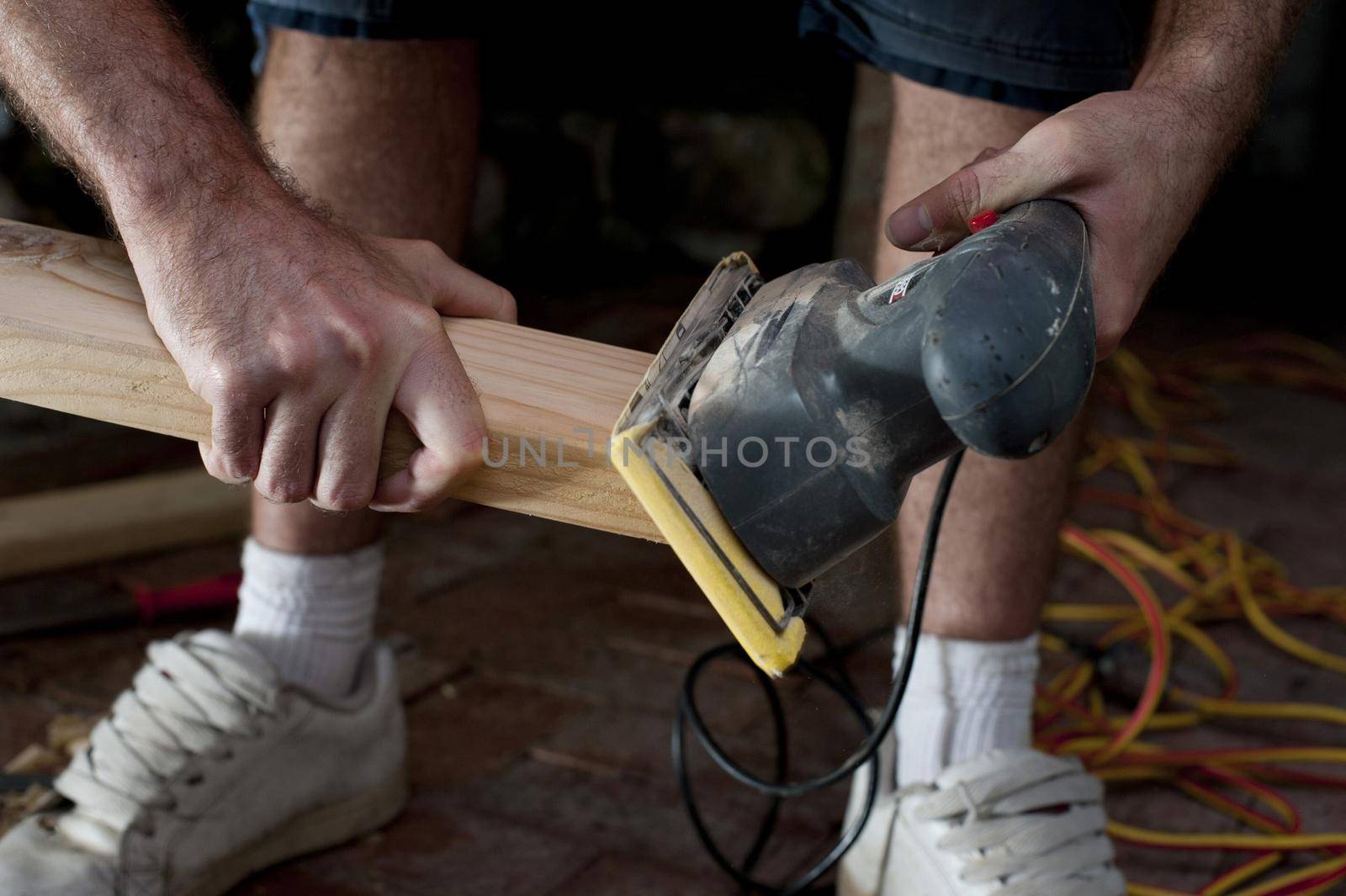 Handyman doing joinery using an electric sander to smooth the surface of a plank of wood before installing it in a house, close up of his hands