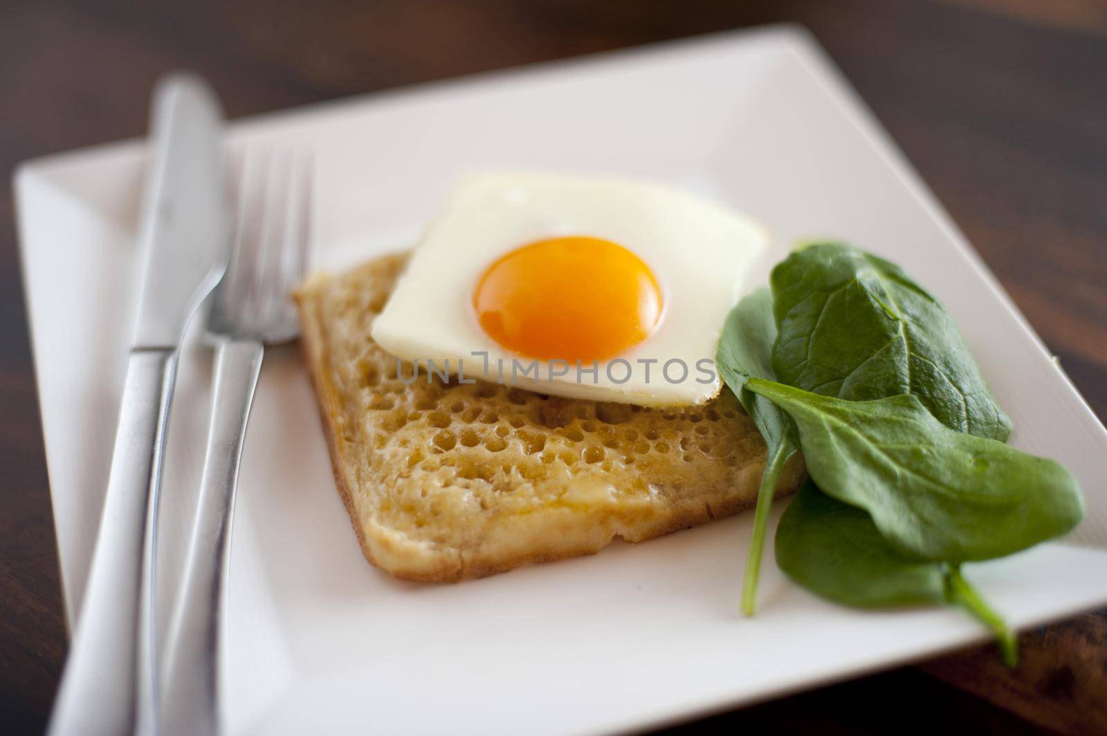 Square fried egg on a square hot buttered crumpet served with fresh baby spinach on a square plate, close up high angle view