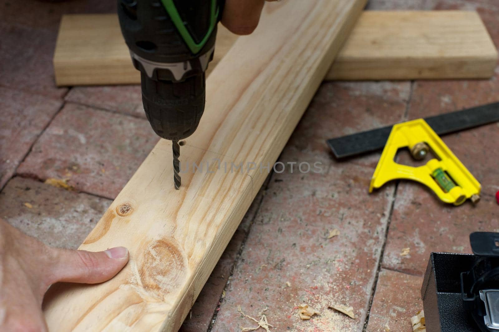 Carpenter drilling a hole in a plank of wood using a handheld electric power drill as he works on a brick floor