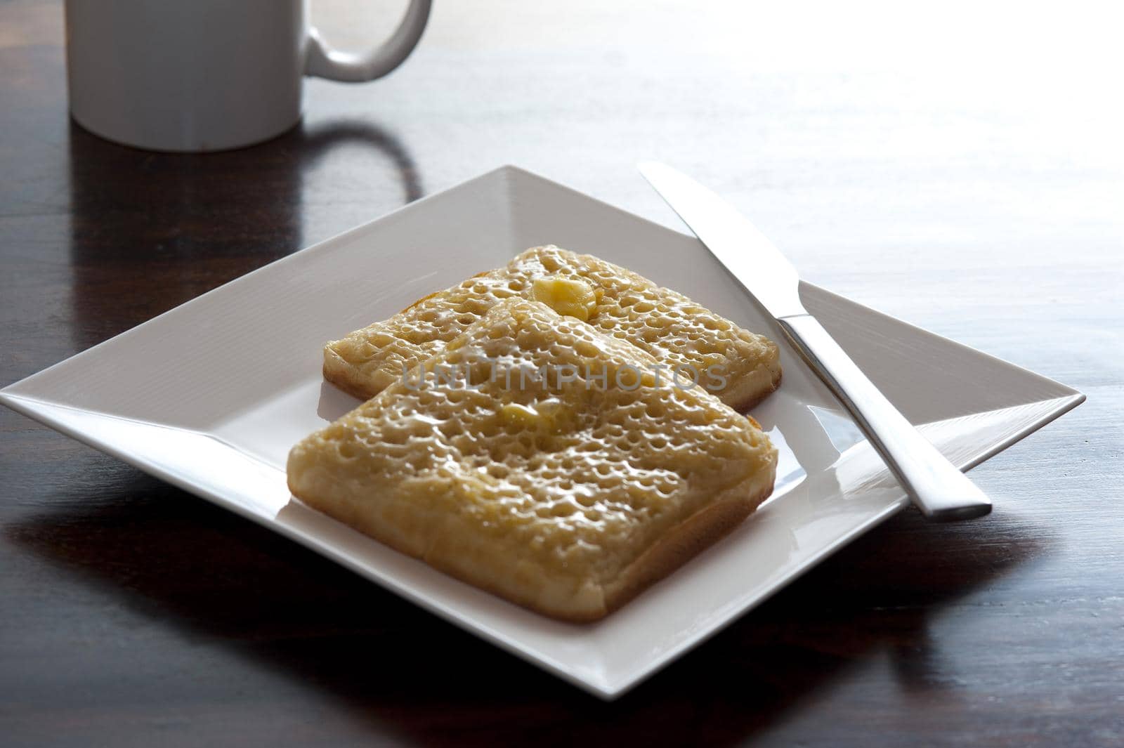 Modern breakfast of two square crumpets with knife on a white plate besides a coffee mug