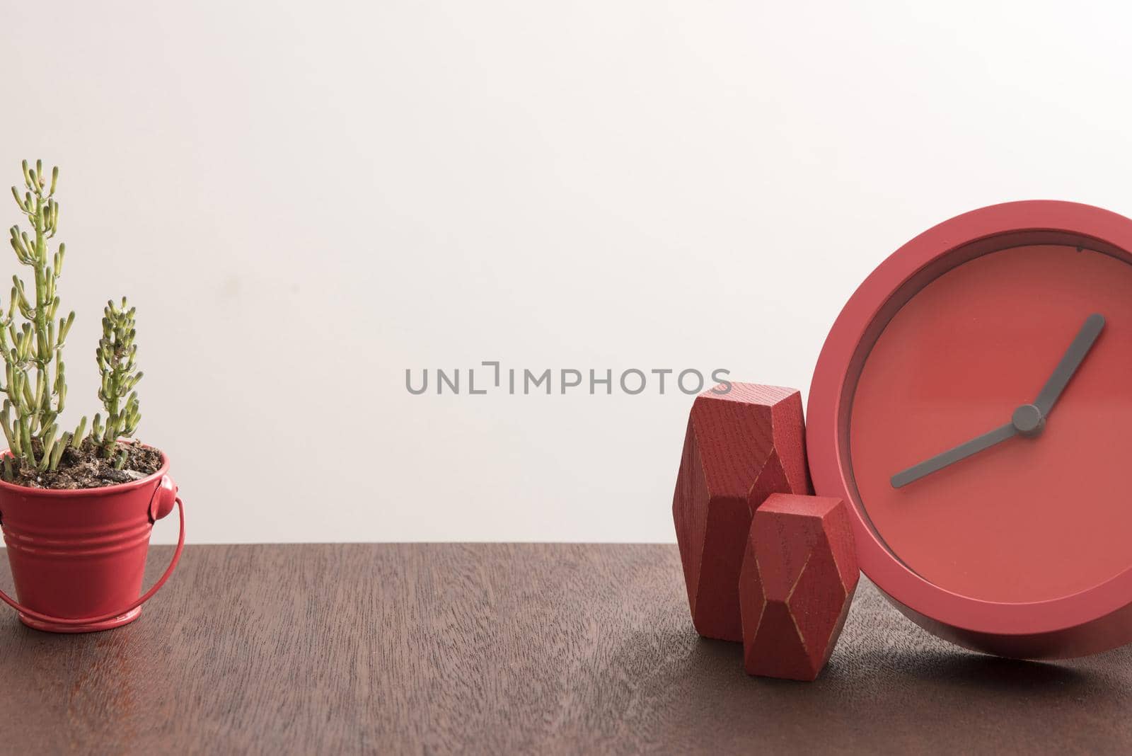 Red faceless clock on beside two polygonal shaped blocks on desk with little plant and copy space