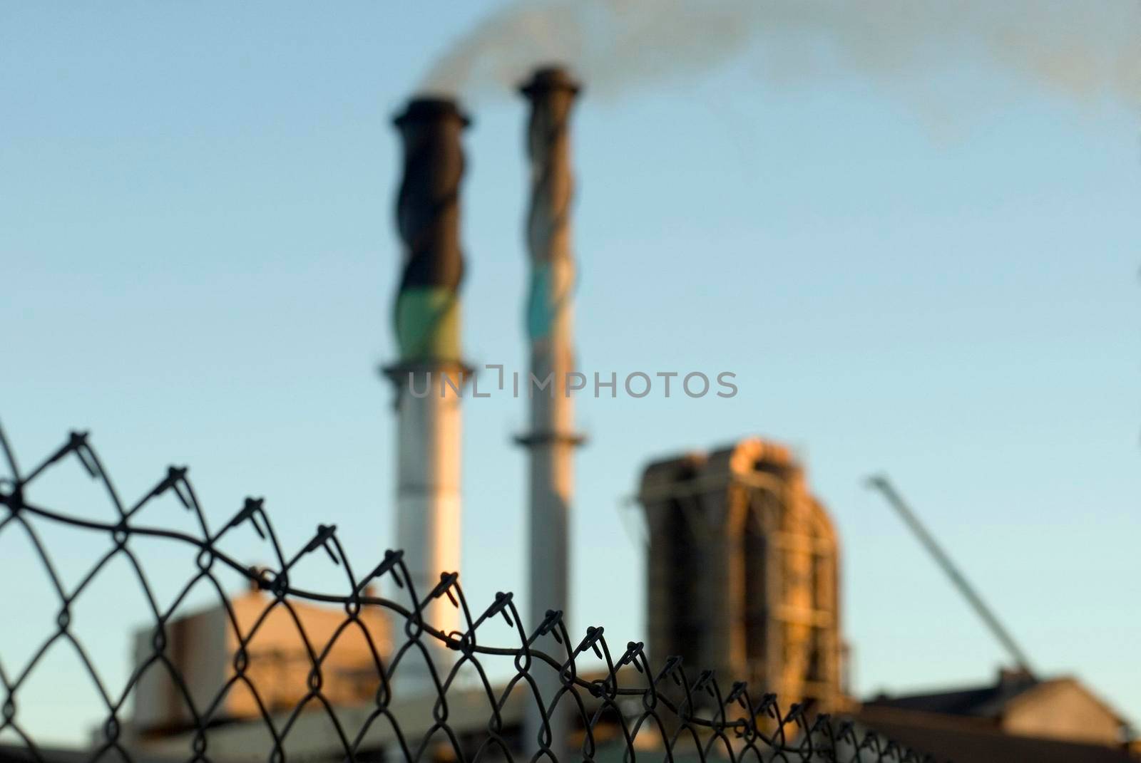 Industrial background of a smoking chimney at an industrial manufacturing plant causing air pollution protected by a wire mesh perimeter fence with focus to the fence