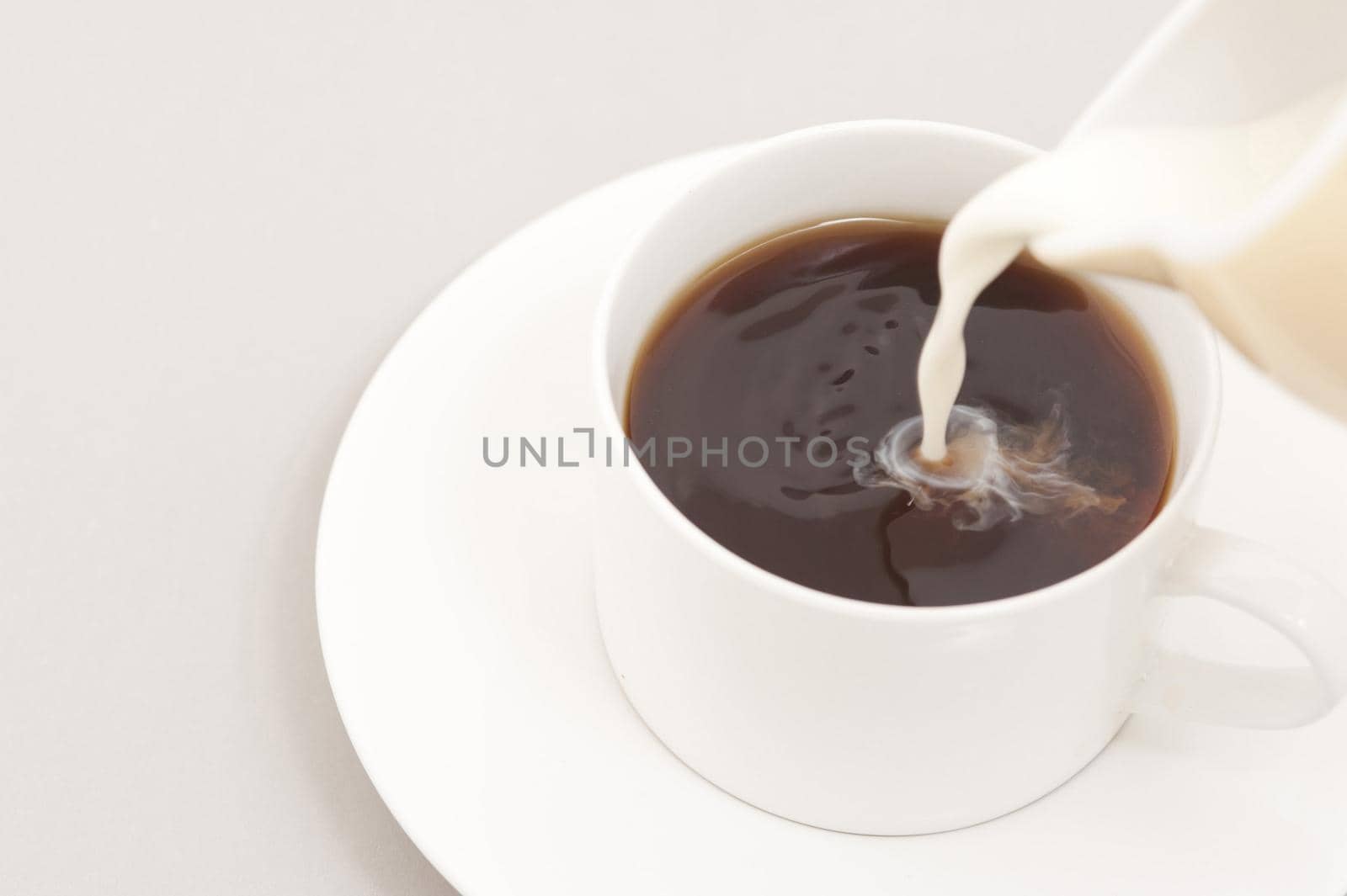 Close up on the spout of a milk jug pouring milk into a cup of black coffee in a generic white cup and saucer