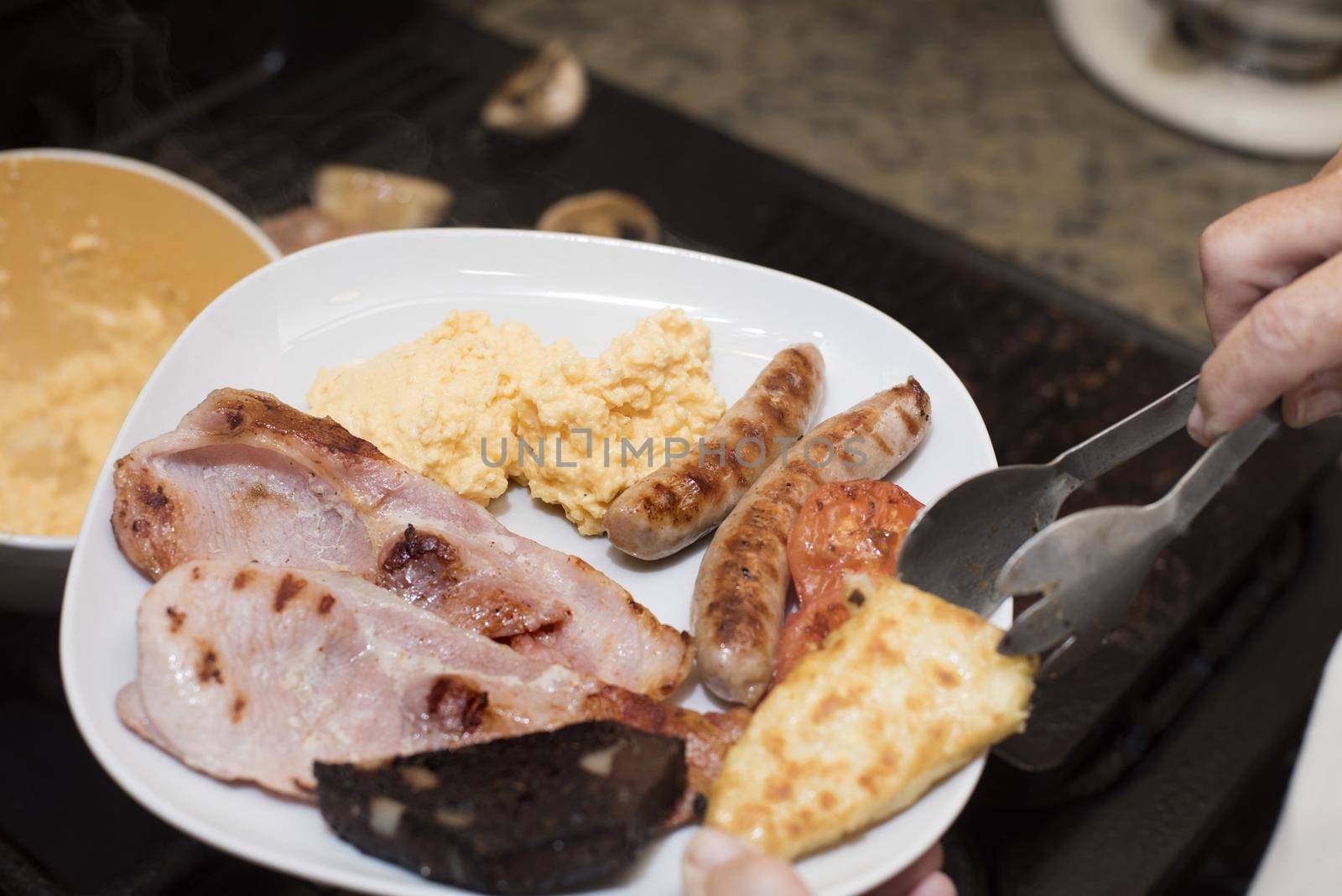 Hand using stainless steel tongs to place a hash brown beside sausage and ham on breakfast plate