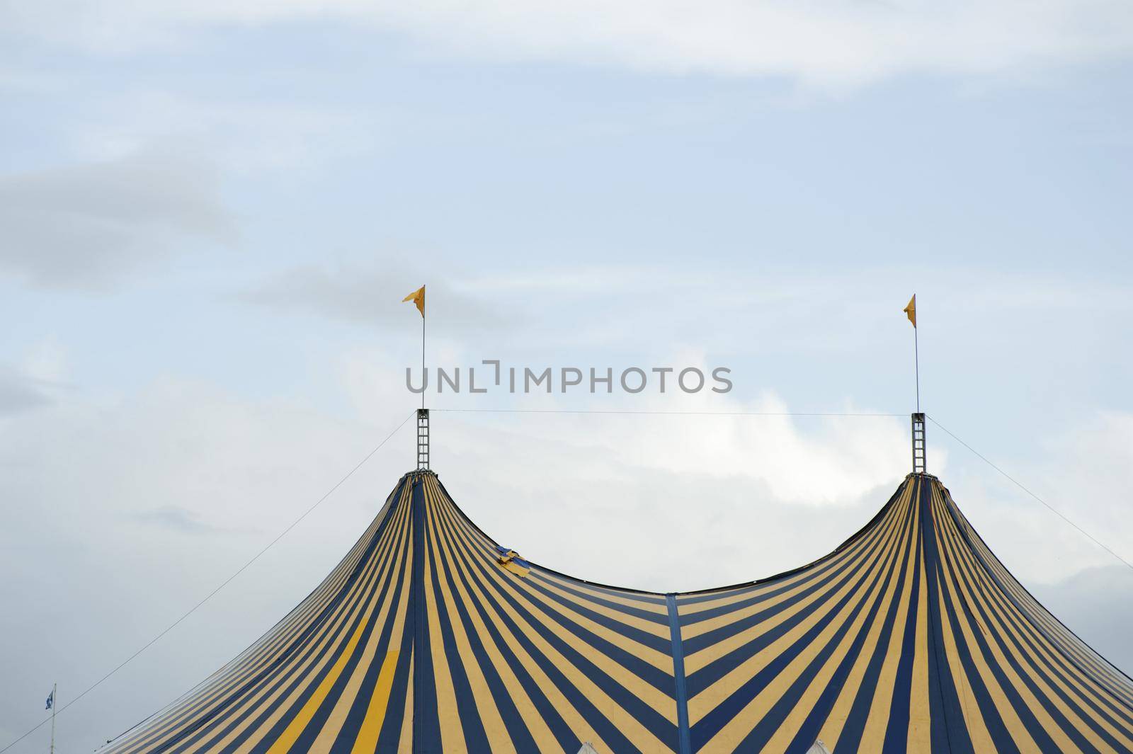 Striped Big Top circus tent with colorful alternating yellow and blue stripes and flags on the poles against a cloudy sky