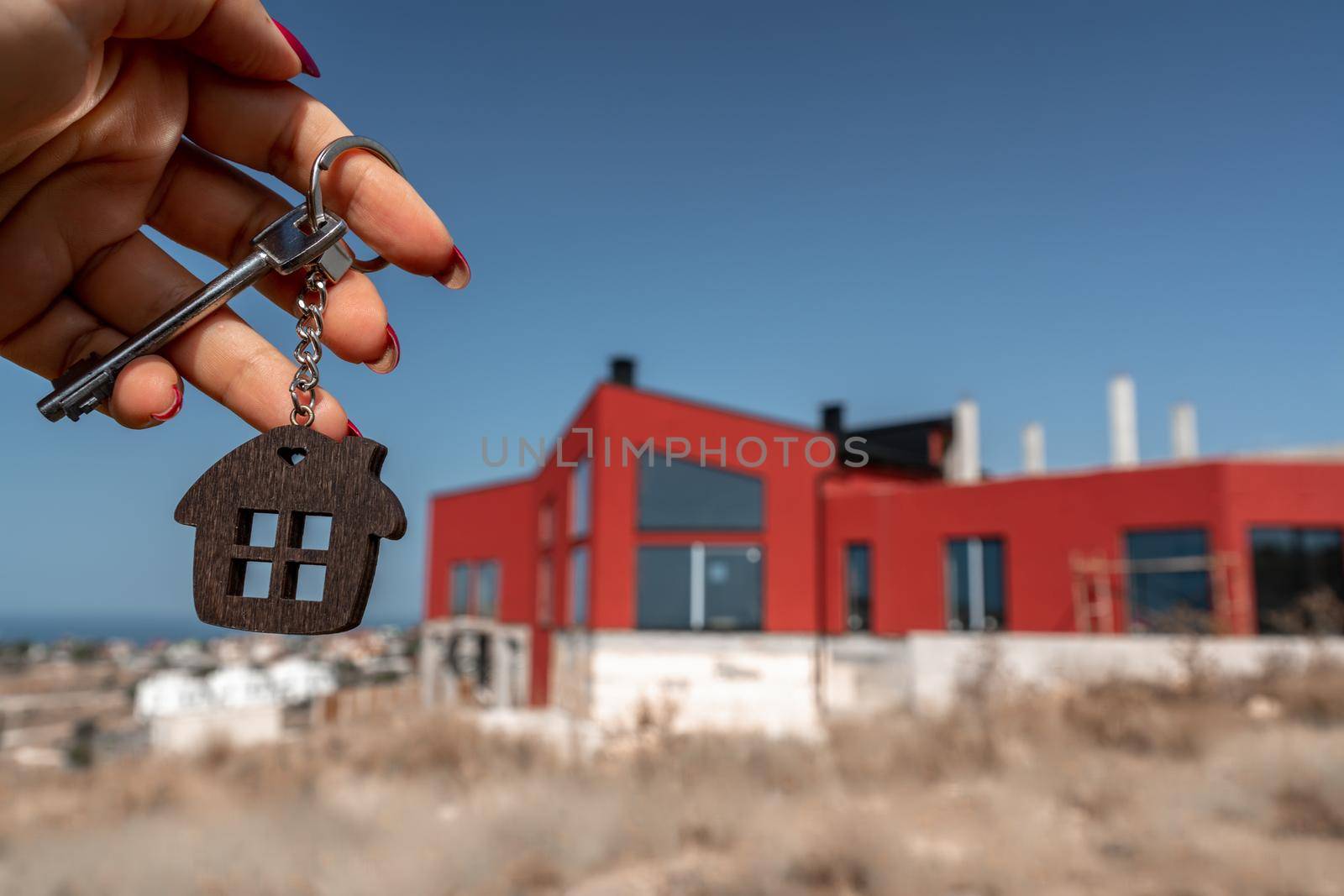 A woman's hand holds a house key against the backdrop of a house under construction. Real estate agent. Buying a house, apartment