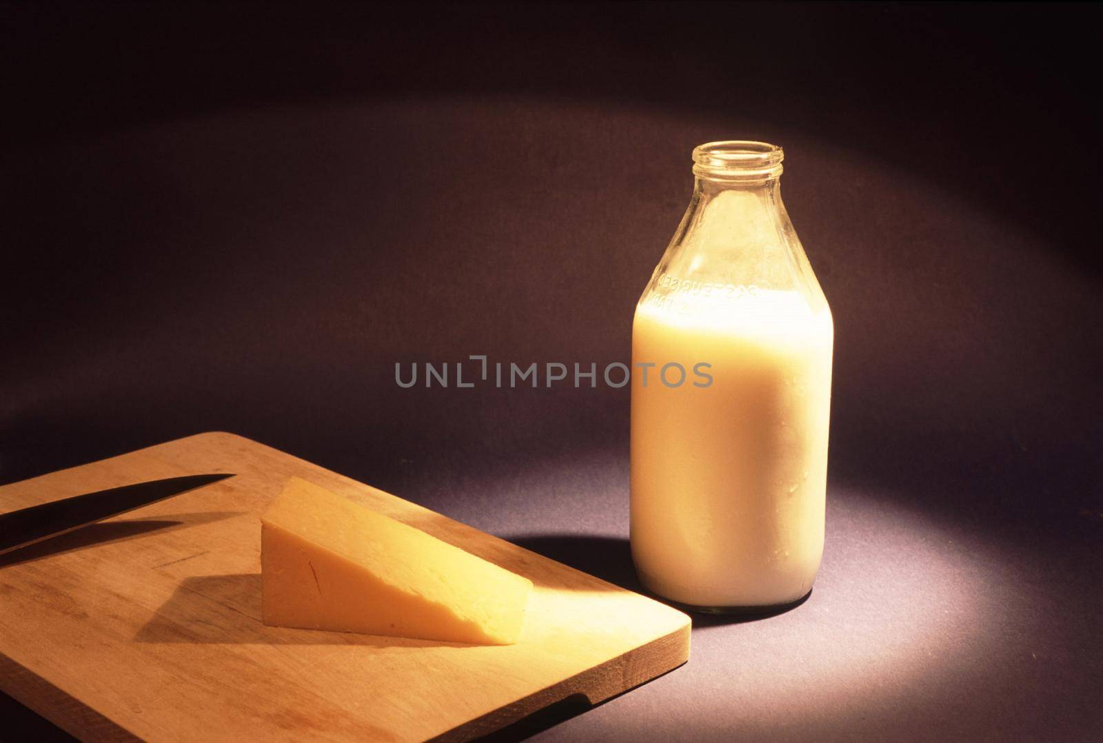 Glass bottle full of fresh creamy cows milk and a wedge of cheddar cheese on a cheeseboard over a dark background with copyspace