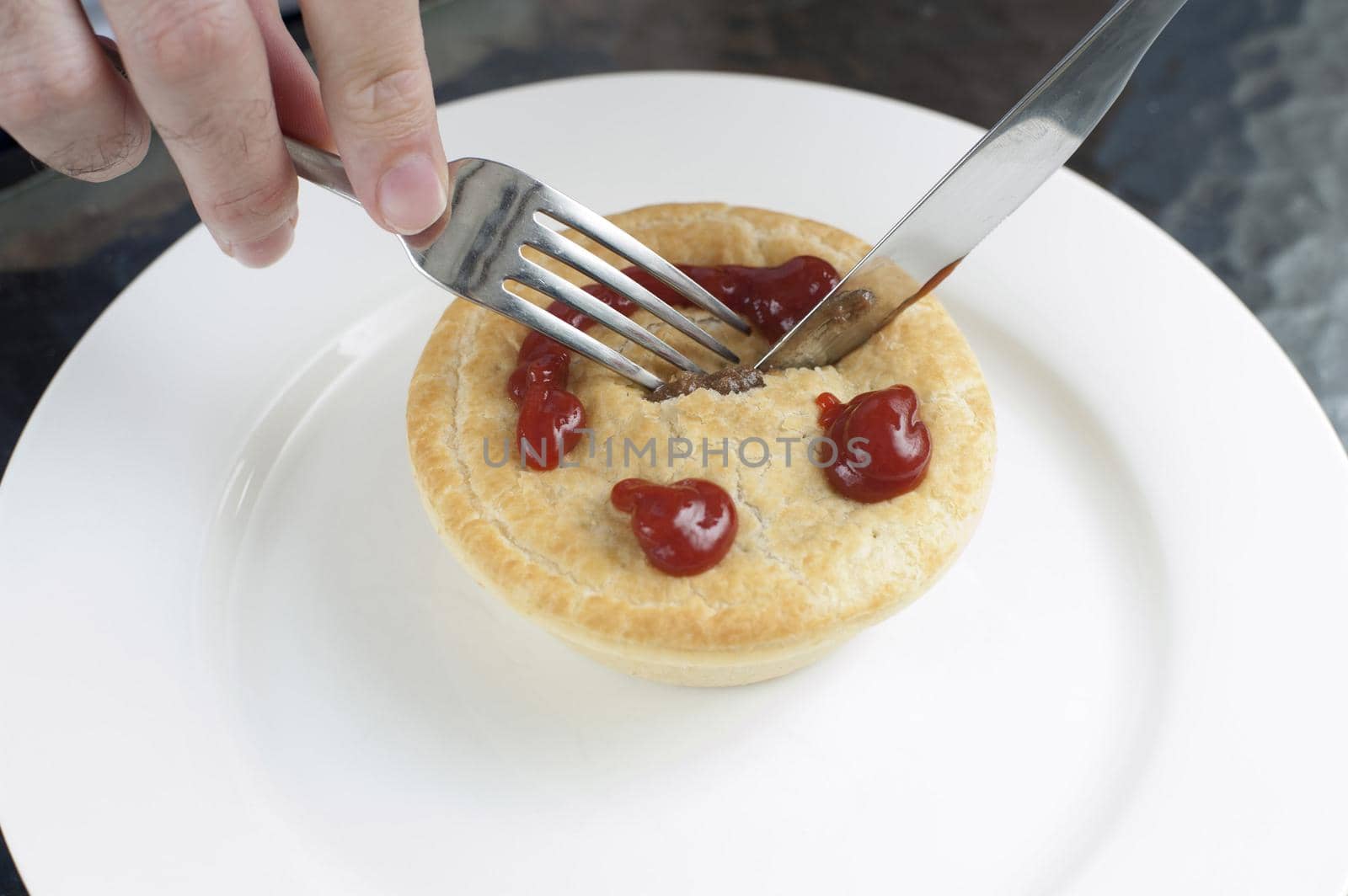 Man eating a freshly baked golden meat pie for lunch with a smiling gravy face on the pastry crust, high angle view as he cuts into it with a knife on a white plate