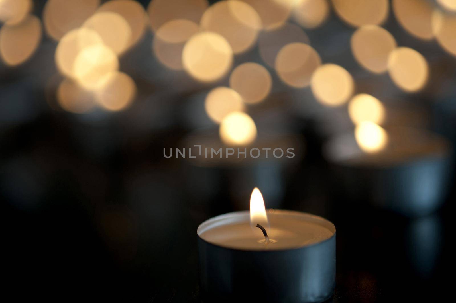 Close-up image of little candle tealight burning in the dark with many glimmering candles blurred in bokeh effect background