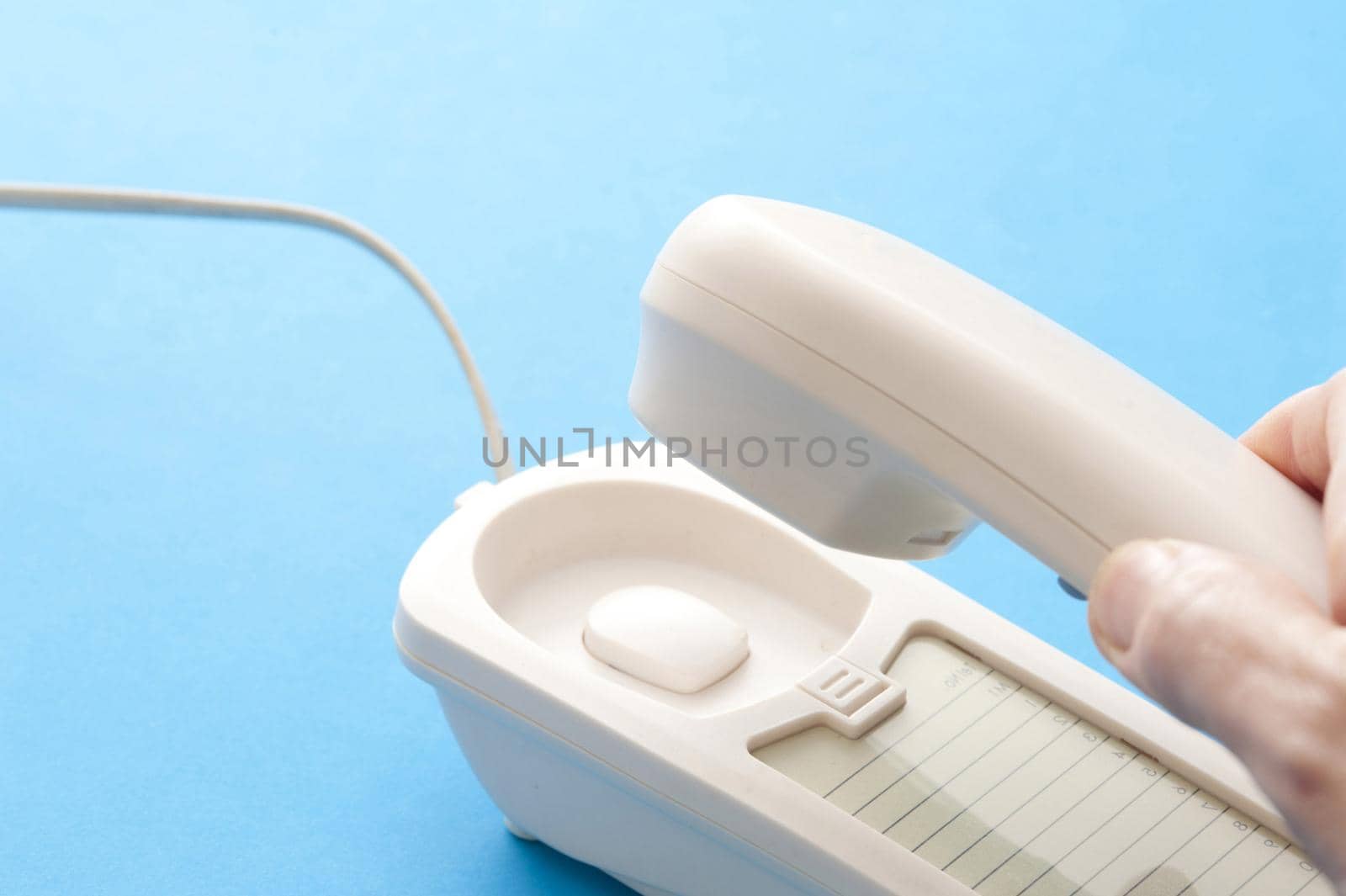 Male hand lifting a telephone handset off the base set of the instrument as he answers a call, close up view over blue