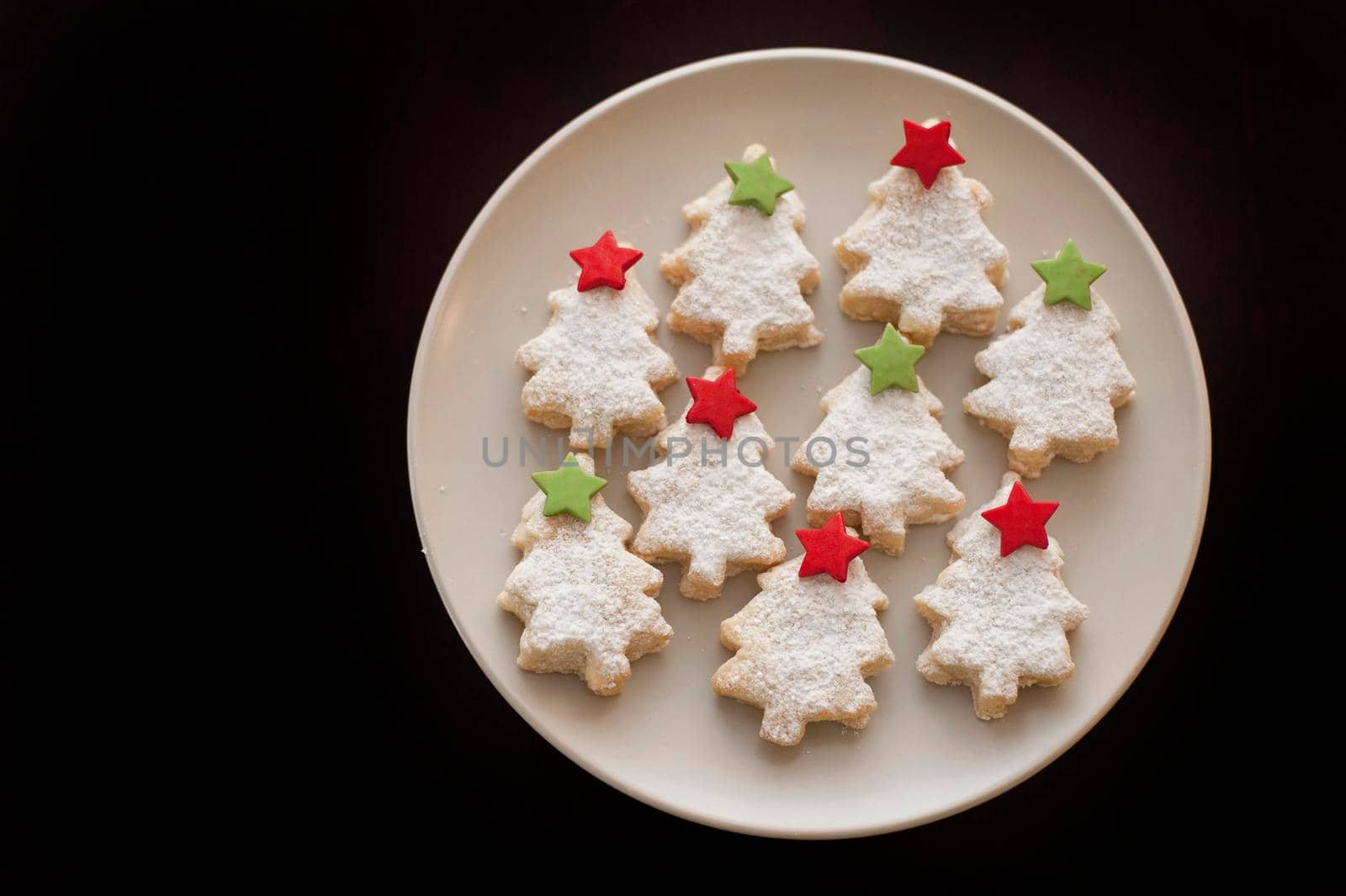 Decorative seasonal Christmas biscuits or cookies in the shape of Christmas trees with colourful stars arranged on a plate on a black background