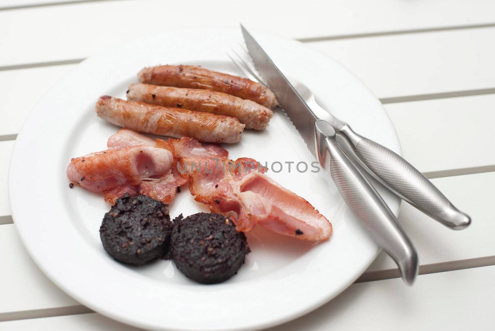 Fried breakfast with black pudding, rashers of crispy bacon and sausages served on a plain white plate