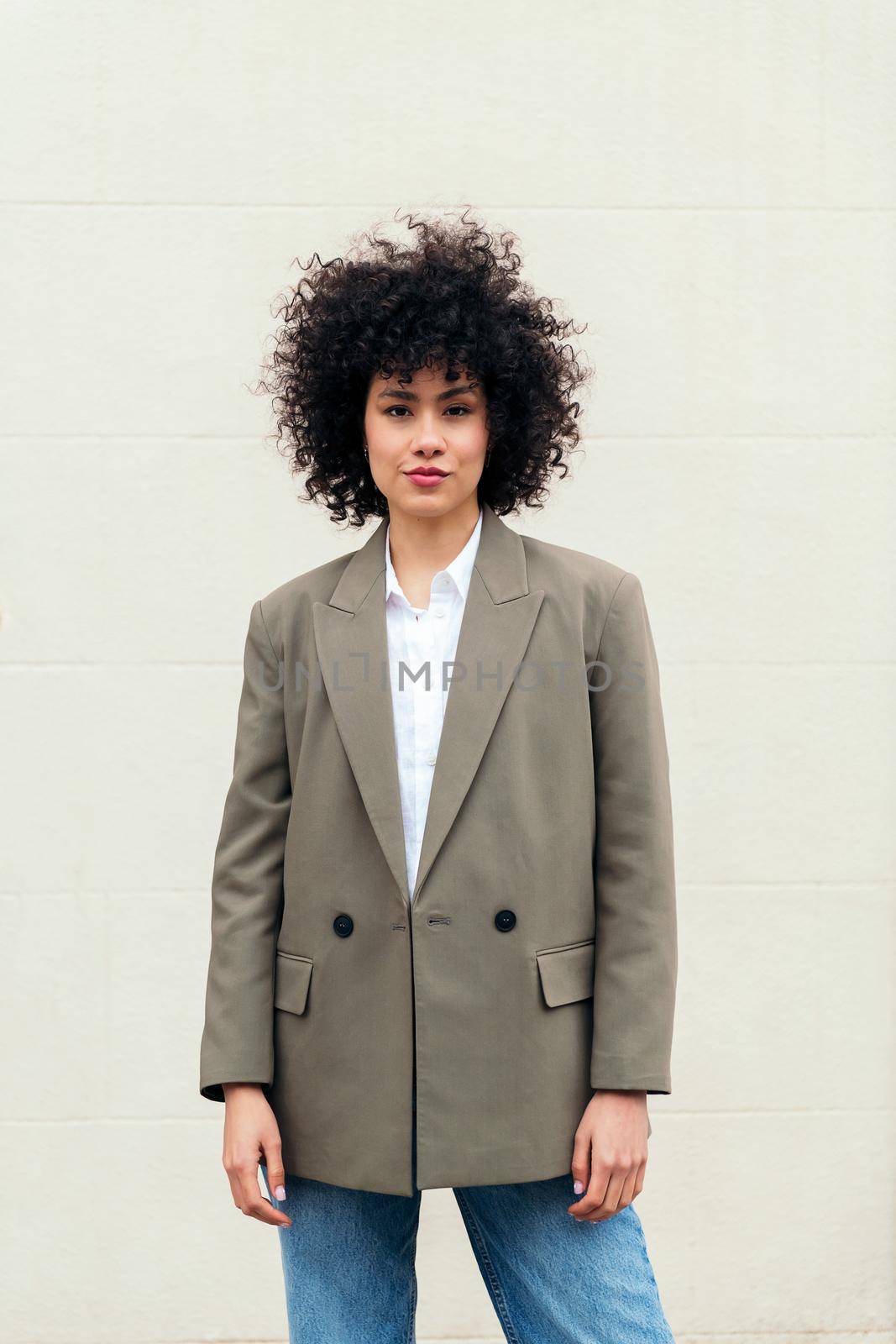 stylish business woman with curly hair wearing a blazer and looking at camera, concept of success and youth