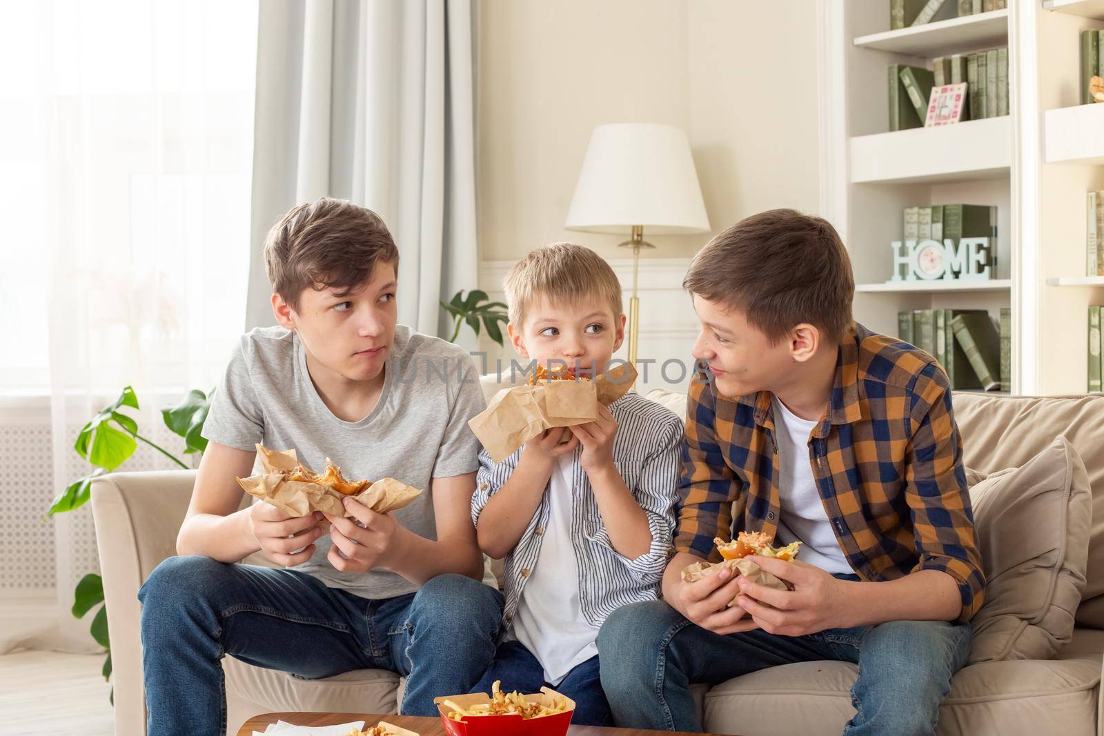 Three happy teenagers, in casual clothes, are sitting on the sofa in the living room, eating fast food, a hamburger, communication