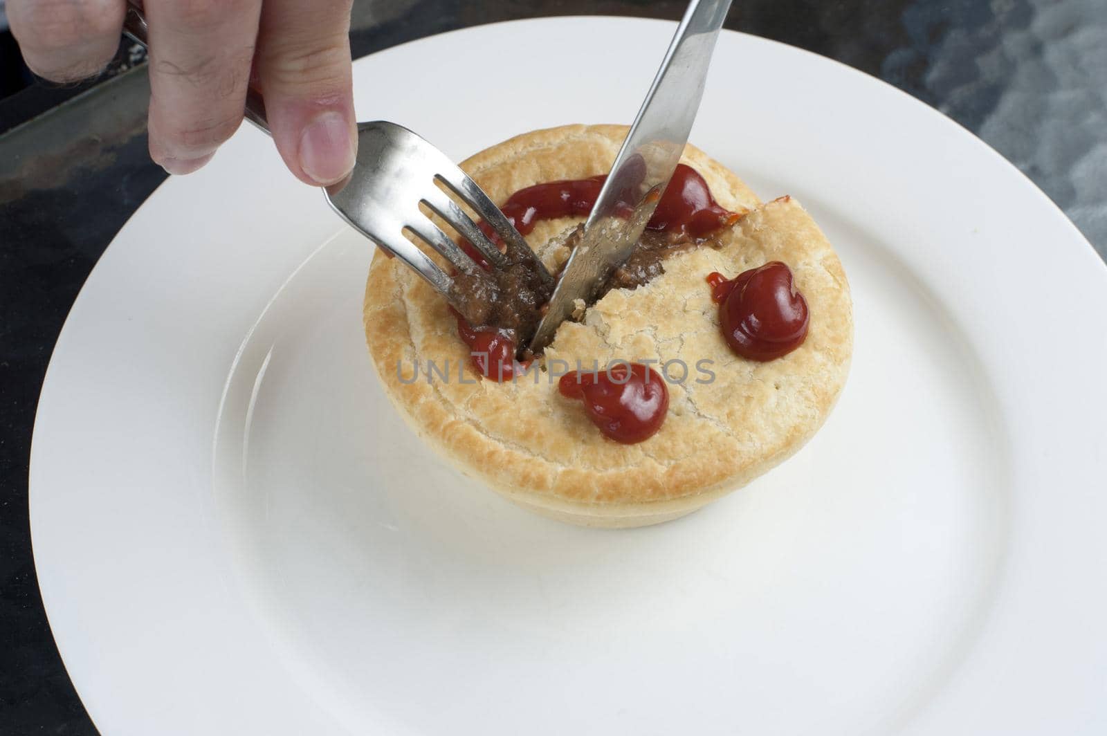 Fresh meat pie with a smiling face formed of sauce on the pastry crust being cut into with a knife and fork for lunch