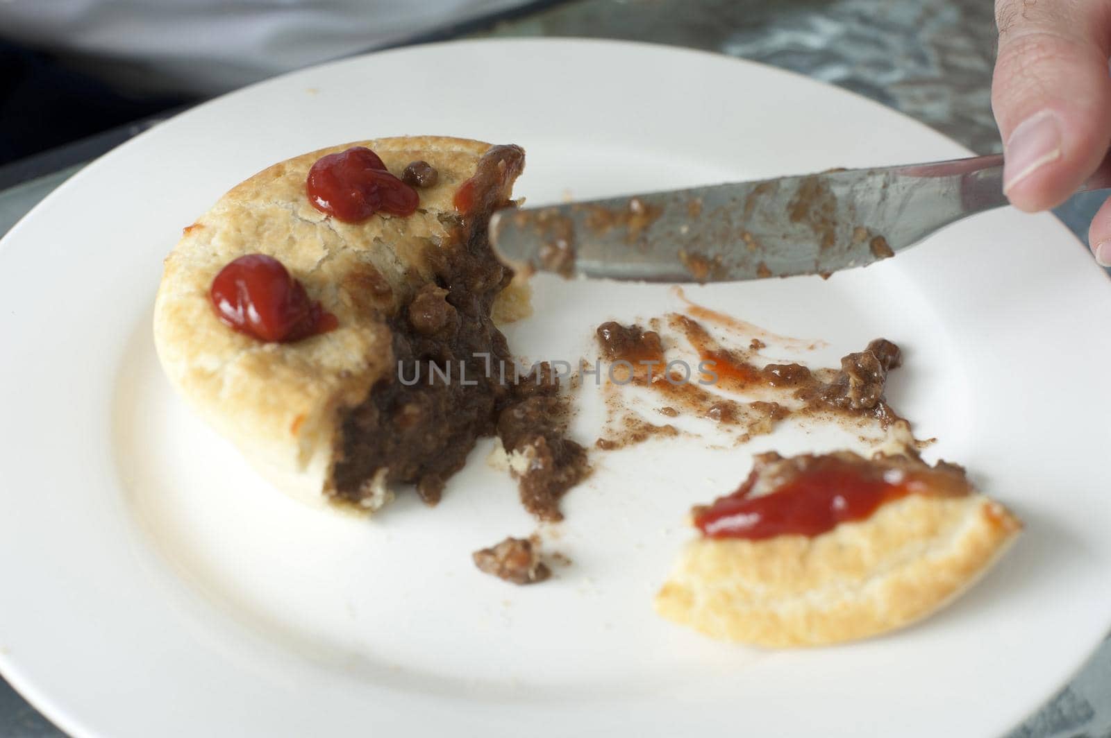 Man eating a pastry meat pie topped with tomato ketchup cutting it with a knife, close up view