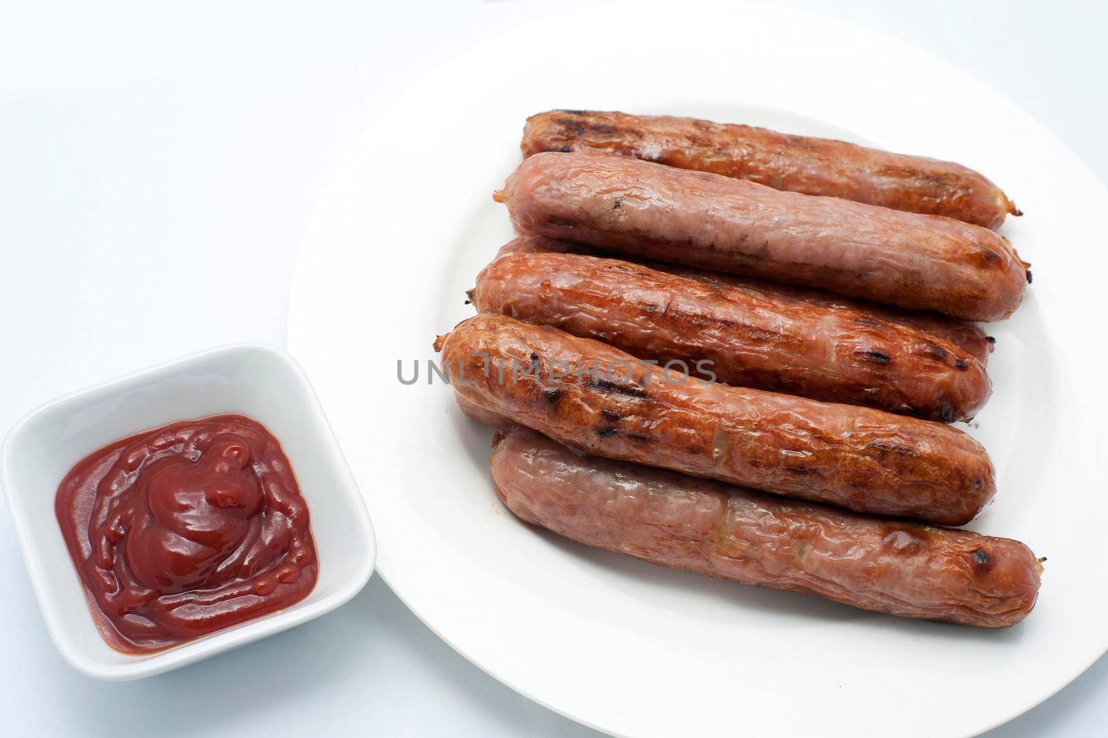 Grilled cooked sausages on a plate with a side dish of tomato ketchup over a white background , high angle view