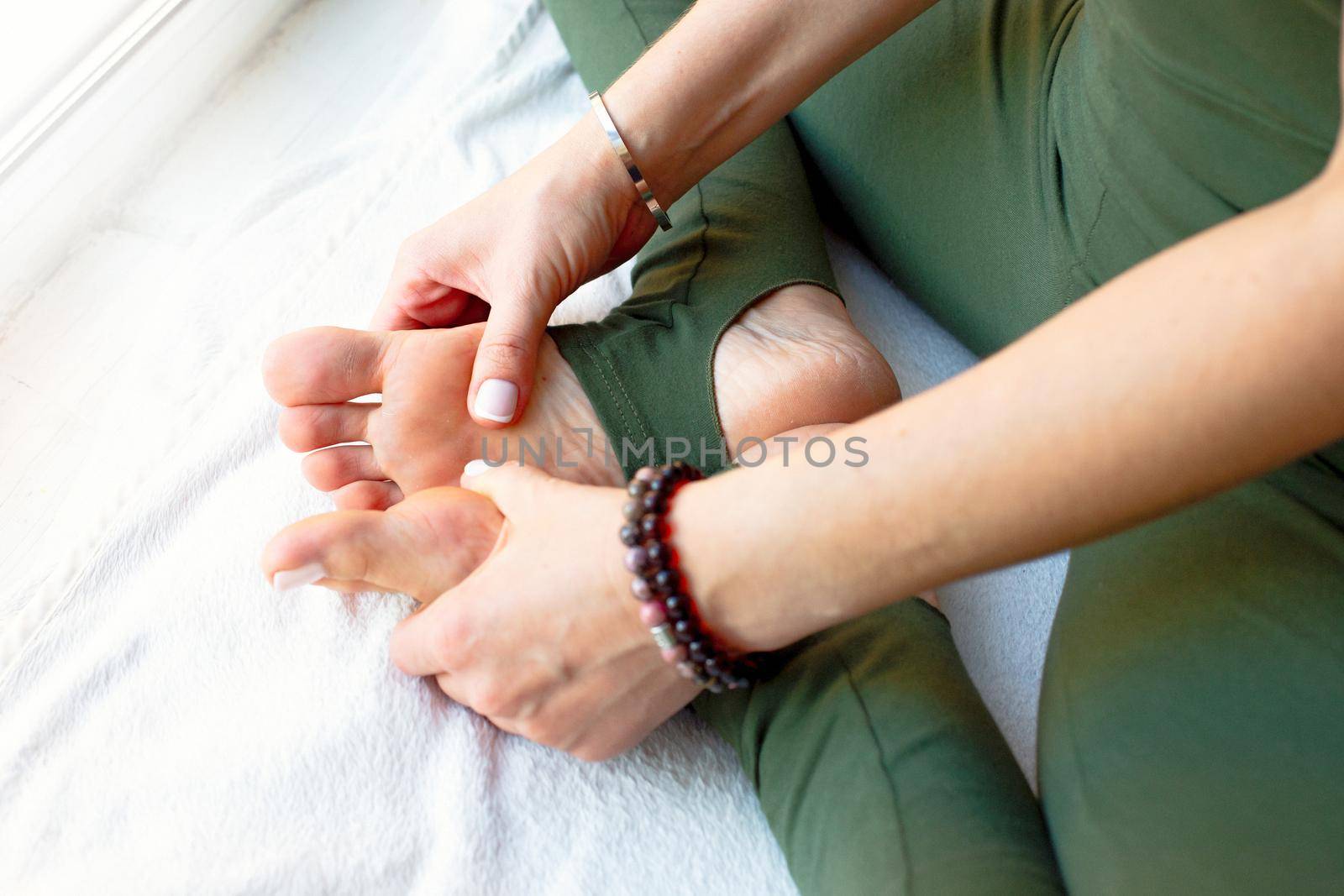 A slender woman in a green jumpsuit, in a yoga pose, clasping her legs with her hands, massaging her feet, sits by a large window. Close-up
