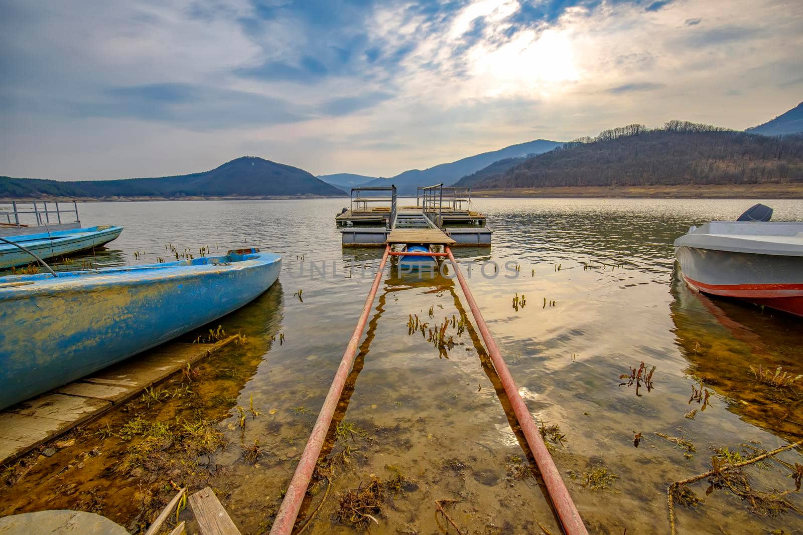 The tranquil afternoon on a lake with a wooden pier and boats in the colorful sky.