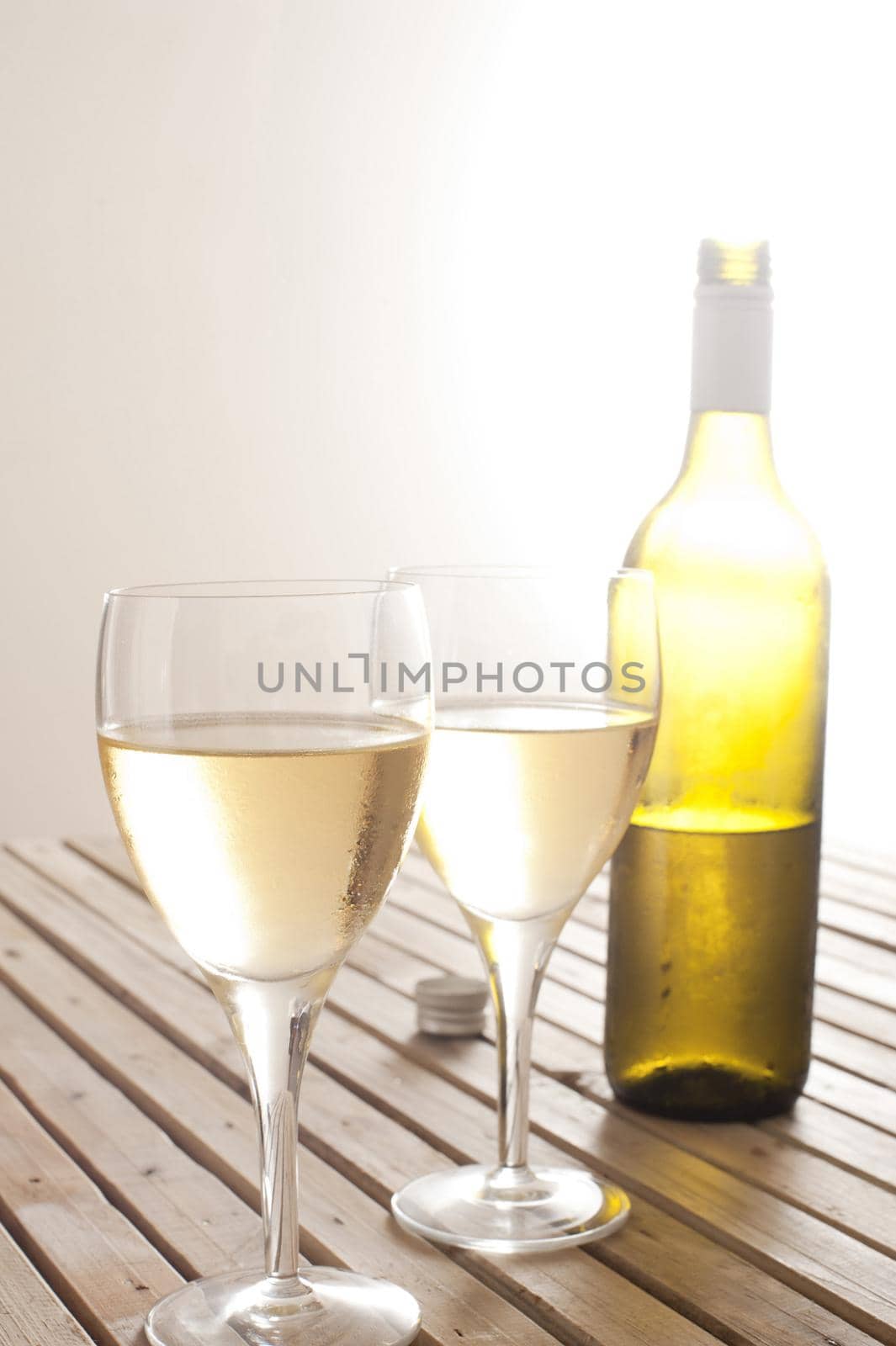 Two glasses of white wine and an unlabelled half full wine bottle standing together on a slatted wooden table with a bright glowing light behind the bottle and copyspace