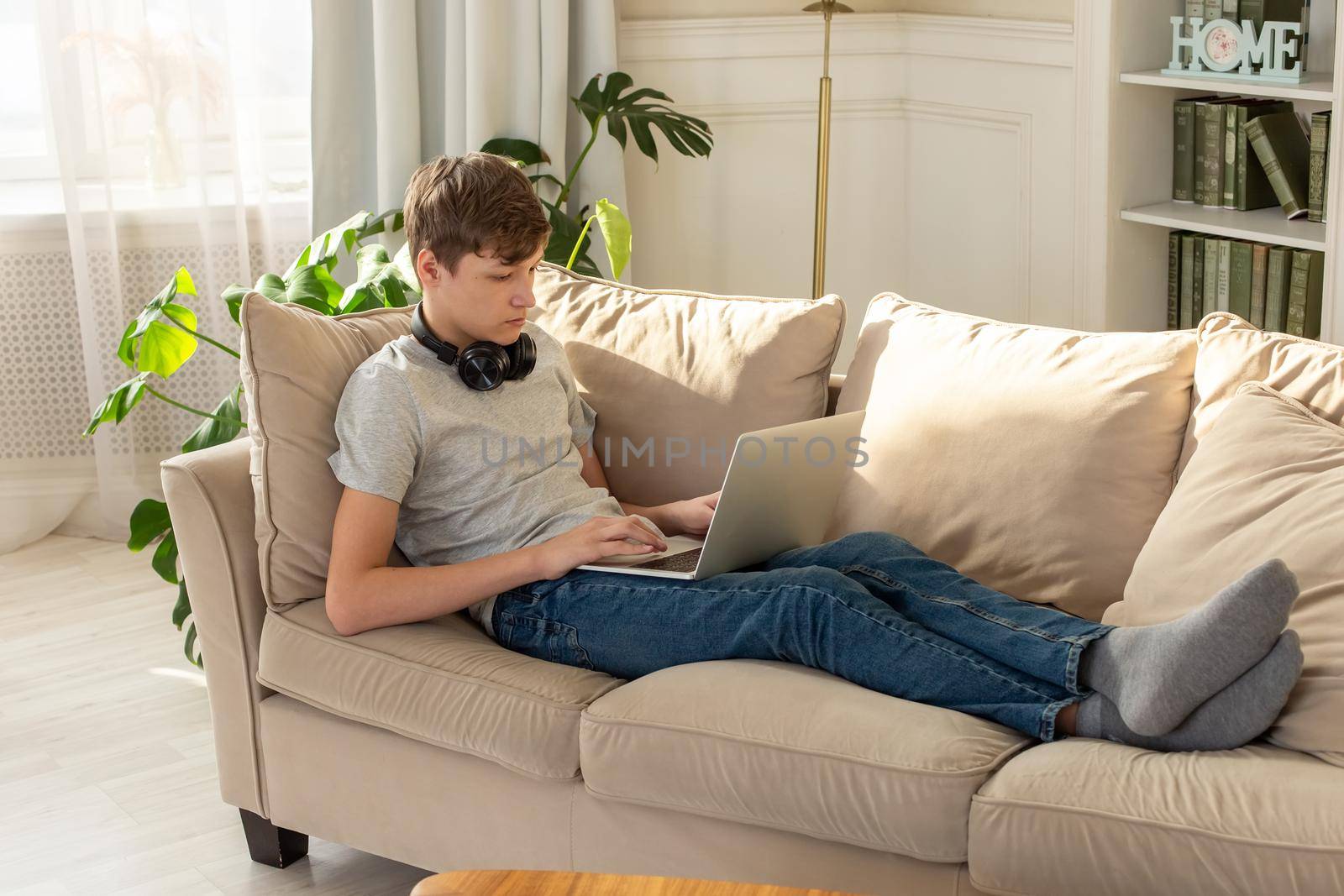 A teenager in a gray T-shirt and blue jeans, lying on a beige sofa, in room with plant, wearing black headphones around his neck, looks into a laptop. Copy space