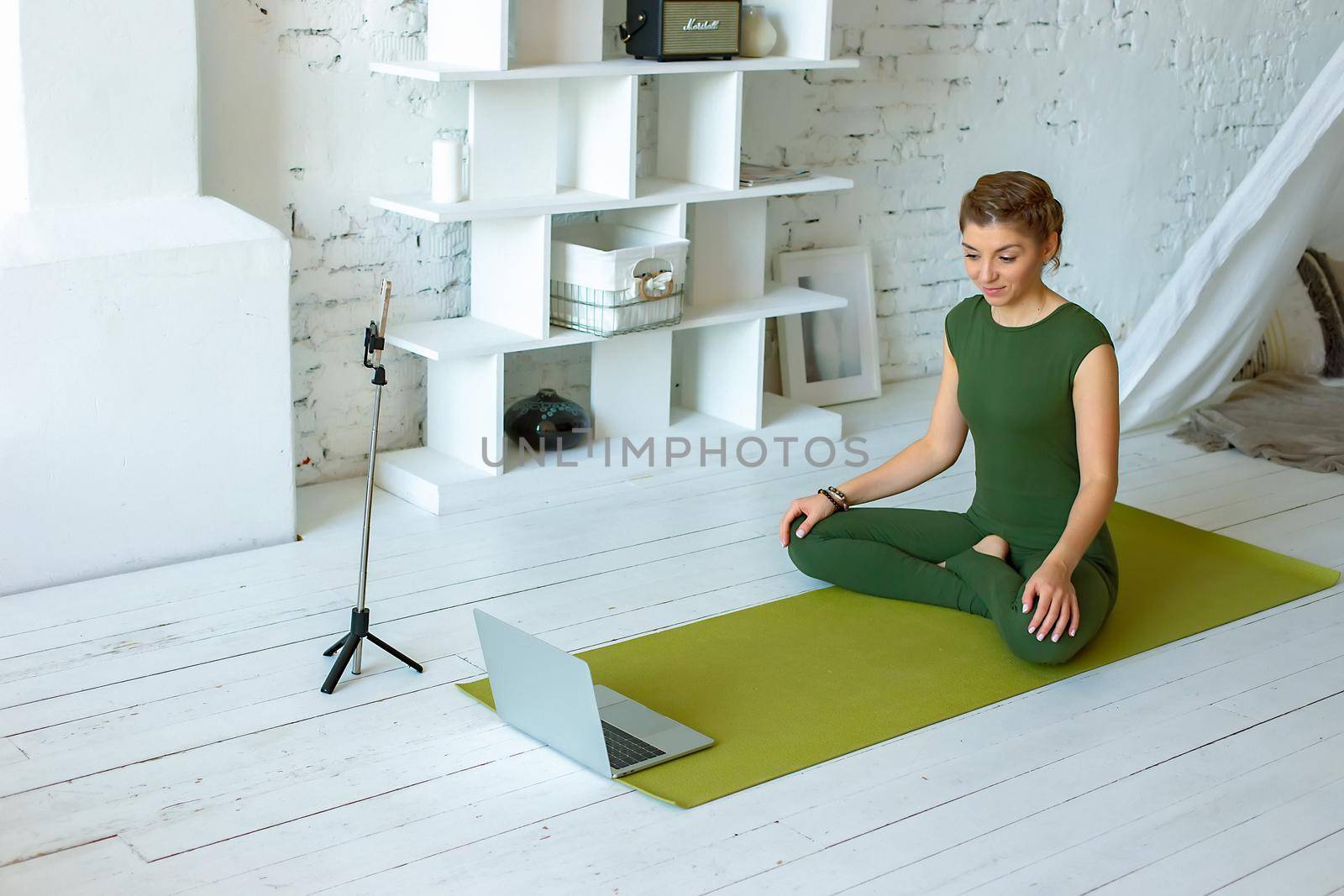 A beautiful and slim woman in green sportswear writes down yoga classes at home, in a light interior, in front of a laptop and a smartphone on a tripod