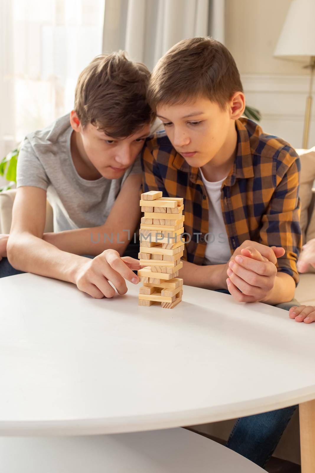 A two teen boys are enthusiastically playing a board game made of wooden rectangular blocks stacked in the form of a tower in a sunny room. Close-up. Vertical