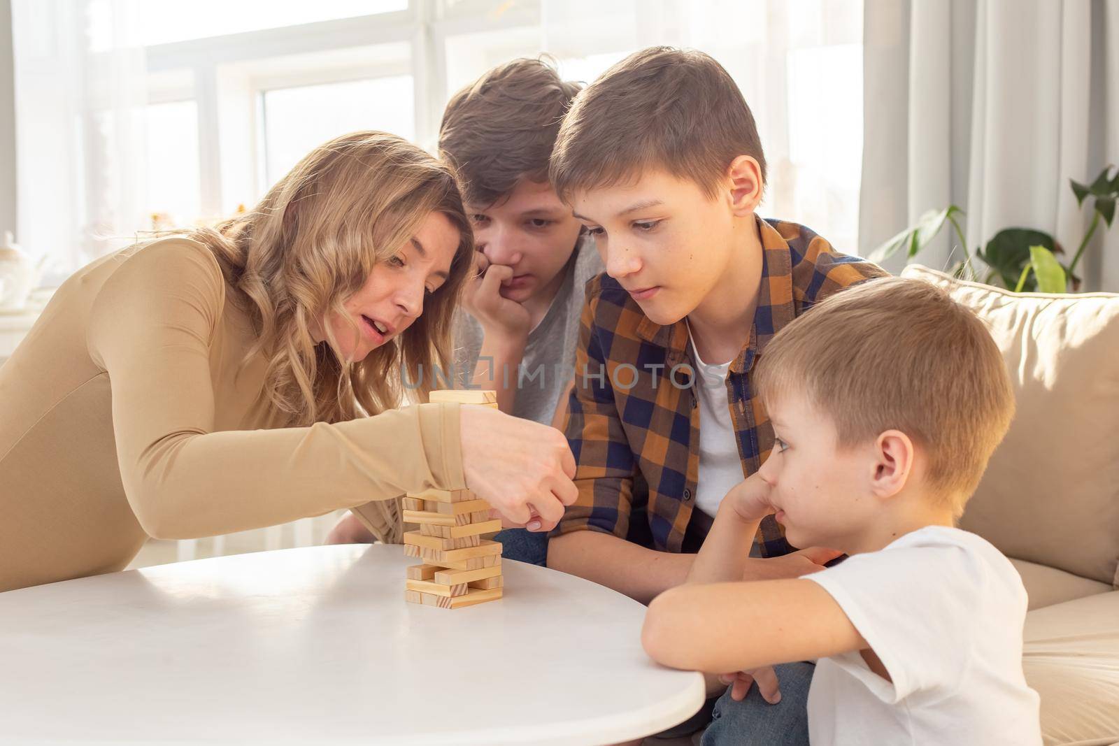 A cute woman and three sons are enthusiastically playing a board game made of wooden rectangular blocks stacked in the form of a tower in a sunny room. Close-up