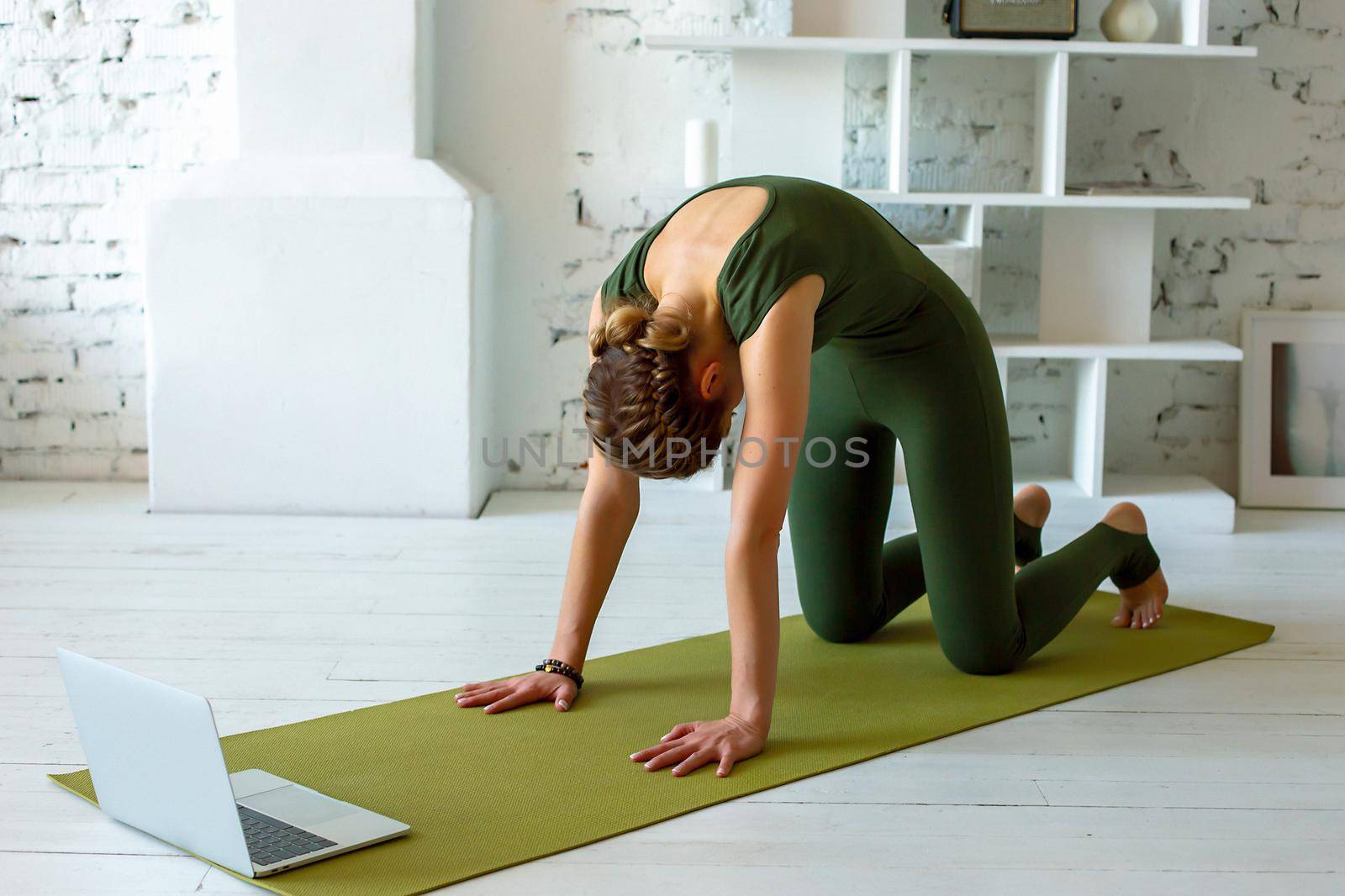 A slender woman in a green sports overalls performs the -cat face down, yoga exercise at home, in a light interior, in front of a laptop