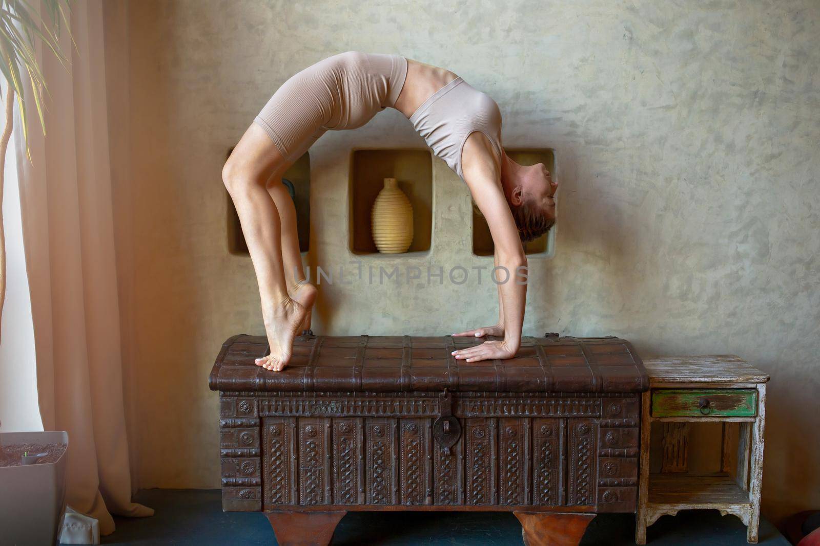 A beautiful and slender woman in a beige top and breeches, stands in a yoga pose, performs the exercise -bridge, in a room with an oriental interior